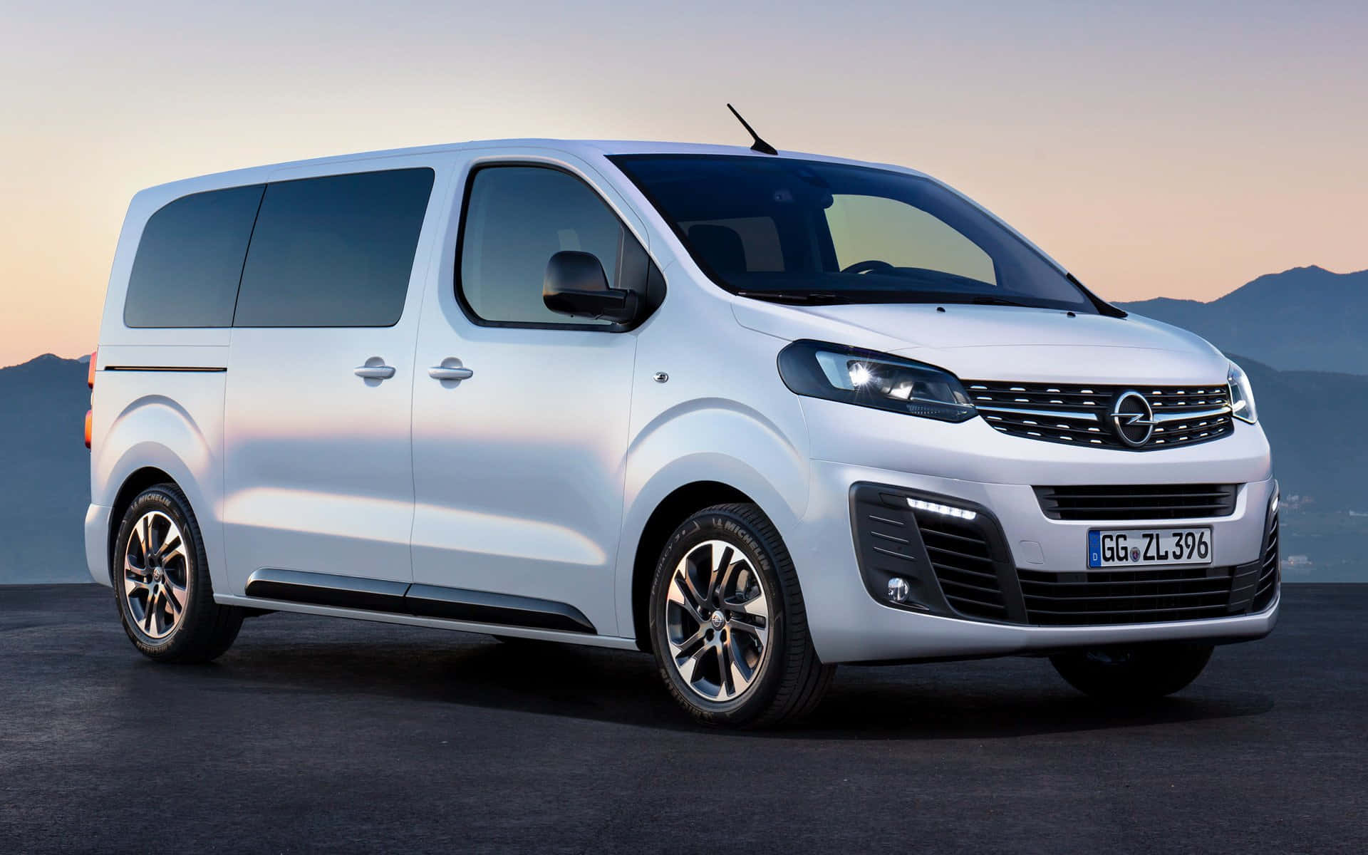 Opel Zafira 桌布 壁紙 — 一輛時尚的白色Opel Vivaro廂型車停在深色地面上，背景有山脈和柔和的日落天空。