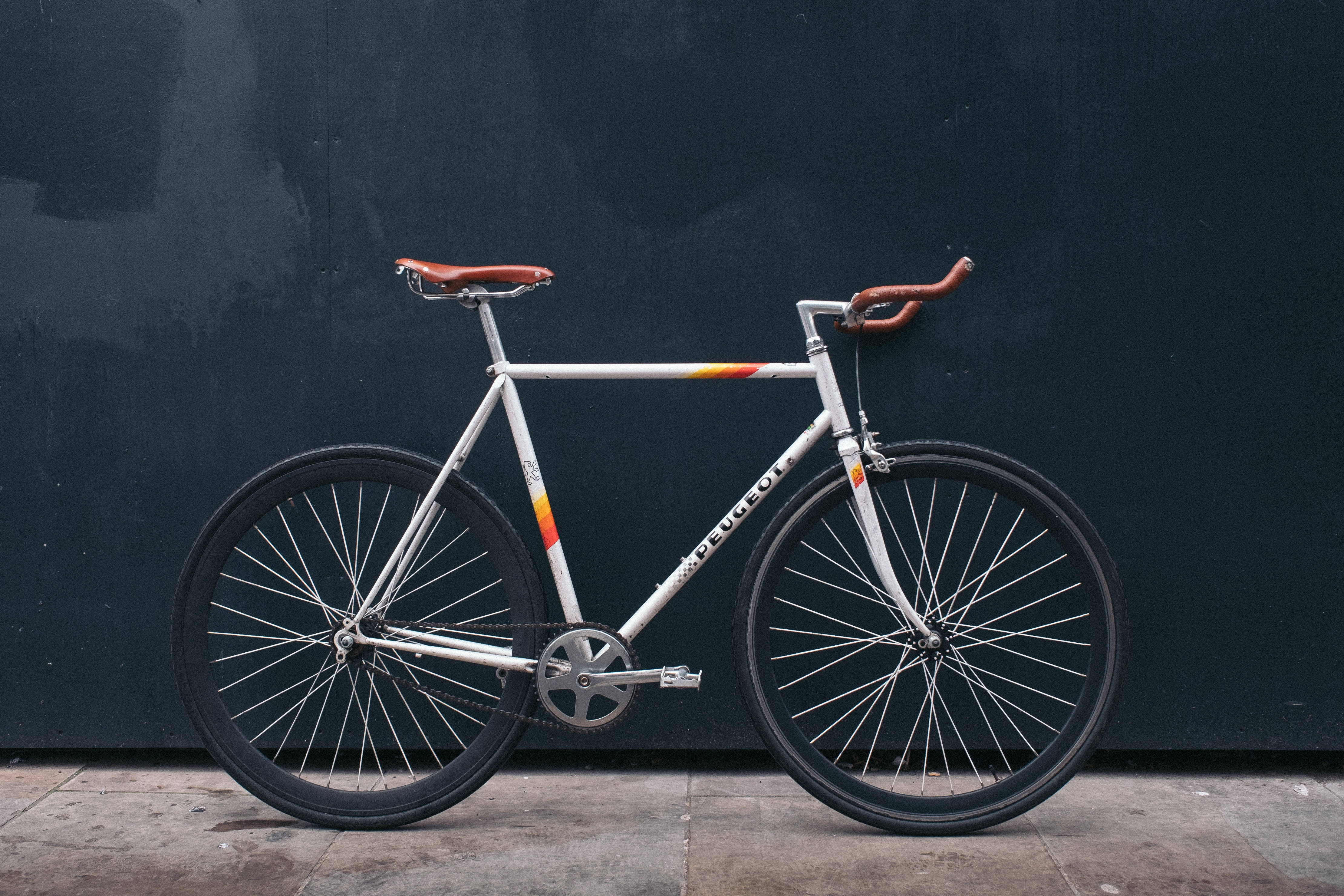 Peugeot 桌布 壁紙 — 一輛時尚的白色Peugeot固定齒輪自行車，帶有棕色皮革裝飾，靠在混凝土人行道上的深色紋理牆上。