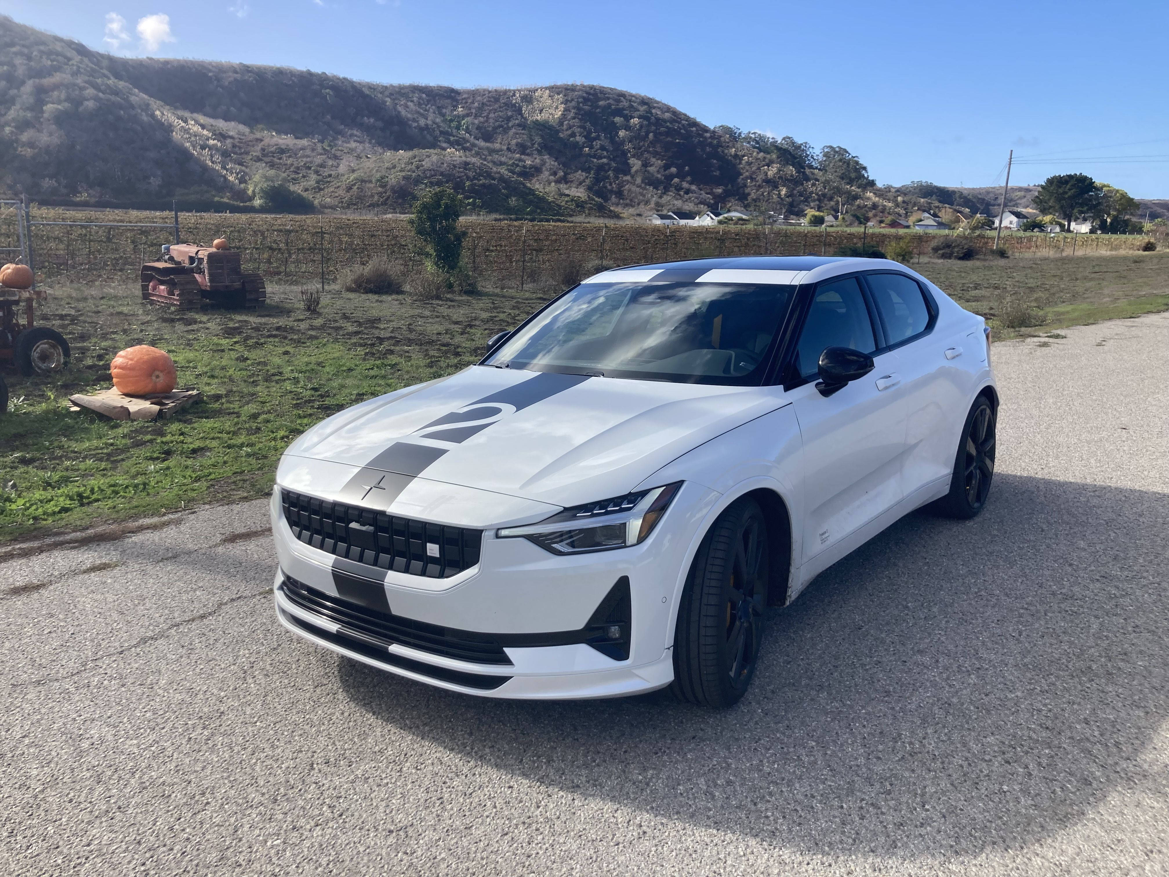 豪華車 桌布 壁紙 — 一輛時尚的白色Polestar 2電動車帶有黑色賽車條紋，停在南瓜和山丘附近的碎石路上，天空湛藍清澈。