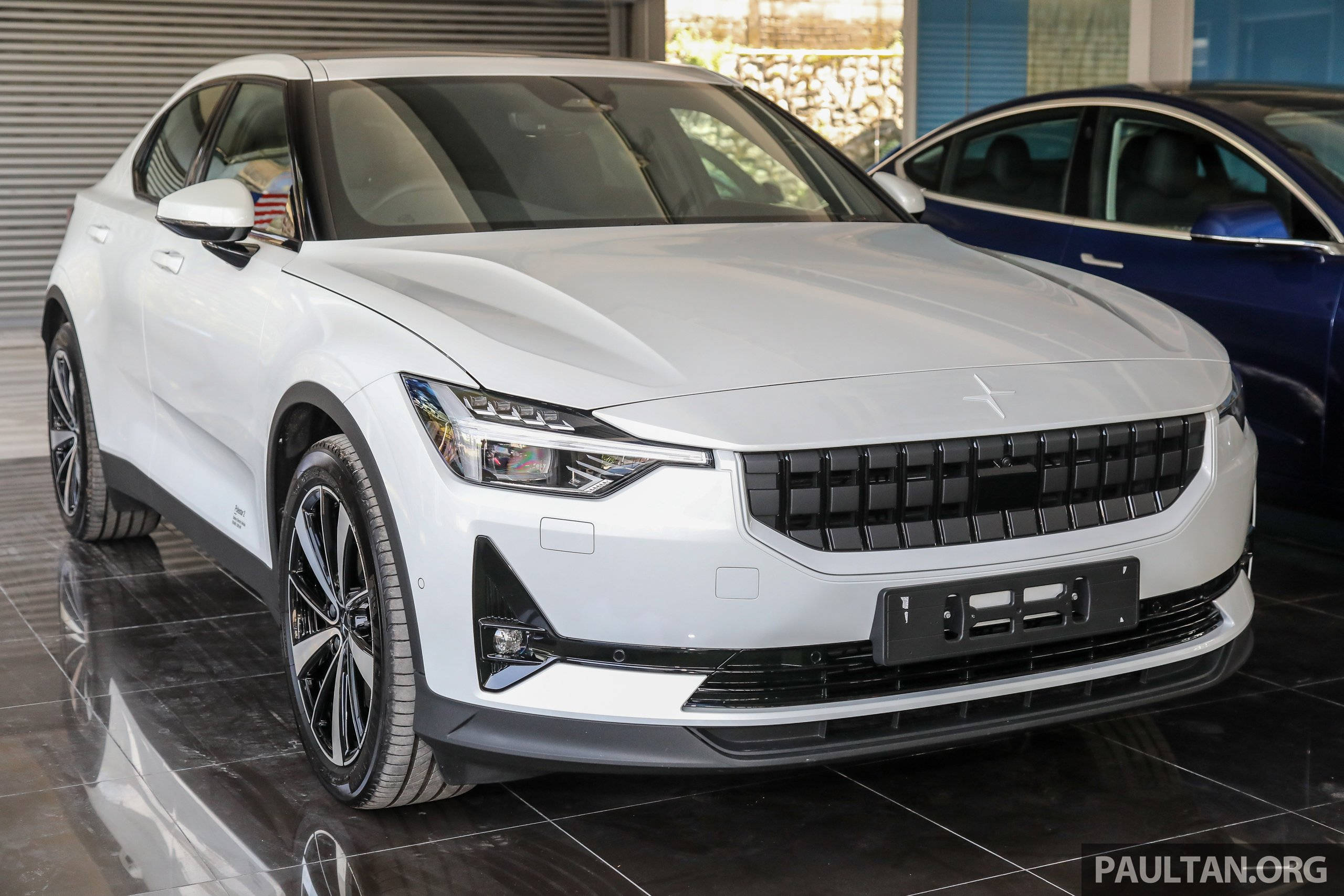 豪華車 桌布 壁紙 — 一輛時尚的白色Polestar 2電動車在展廳燈光下閃閃發光，後面可見一輛深藍色轎車。