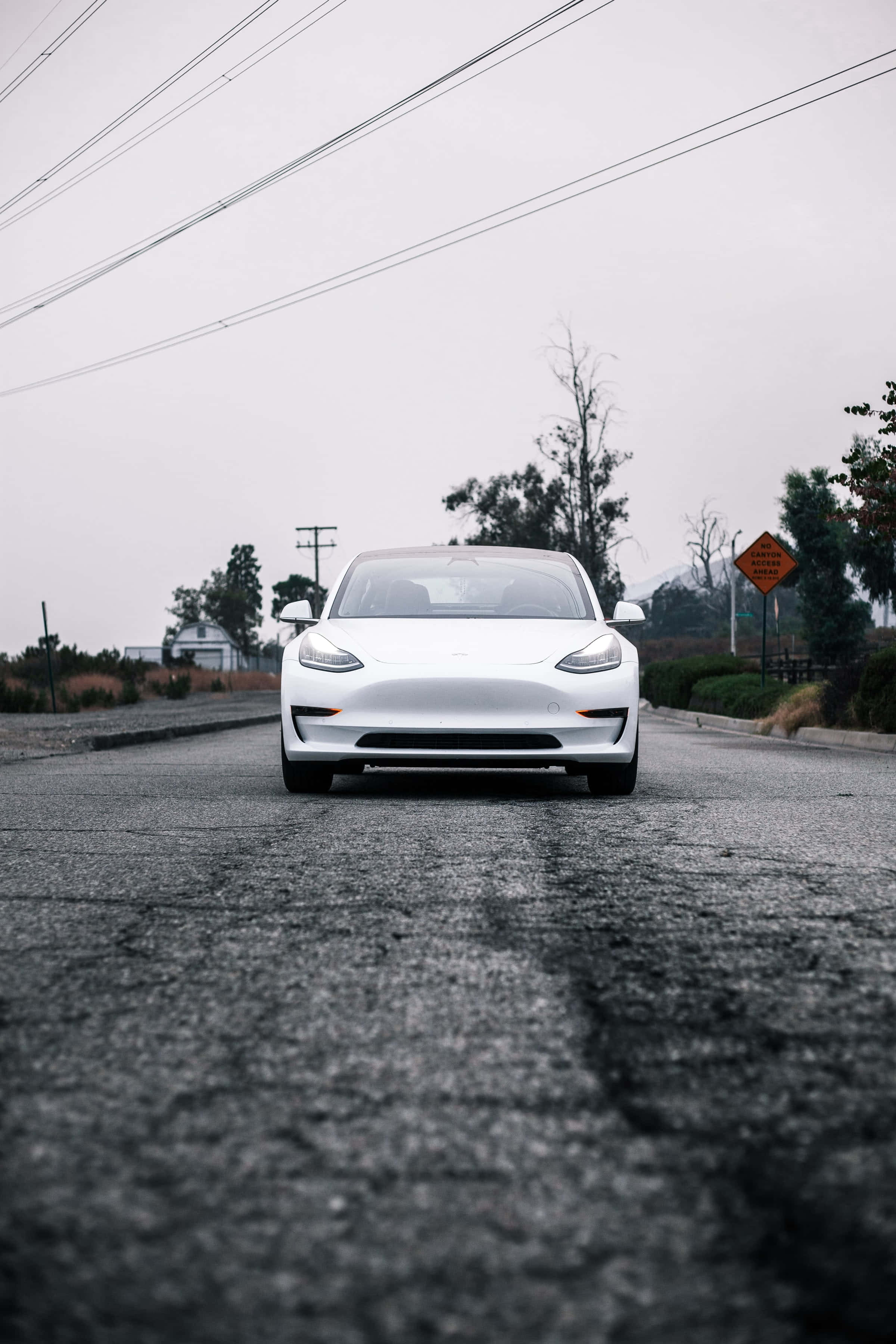 Tesla iPhone 桌布 壁紙 — 一輛時尚的白色Tesla Model 3在潮濕的瀝青路上向前行駛，天空多雲，頭頂有電線。