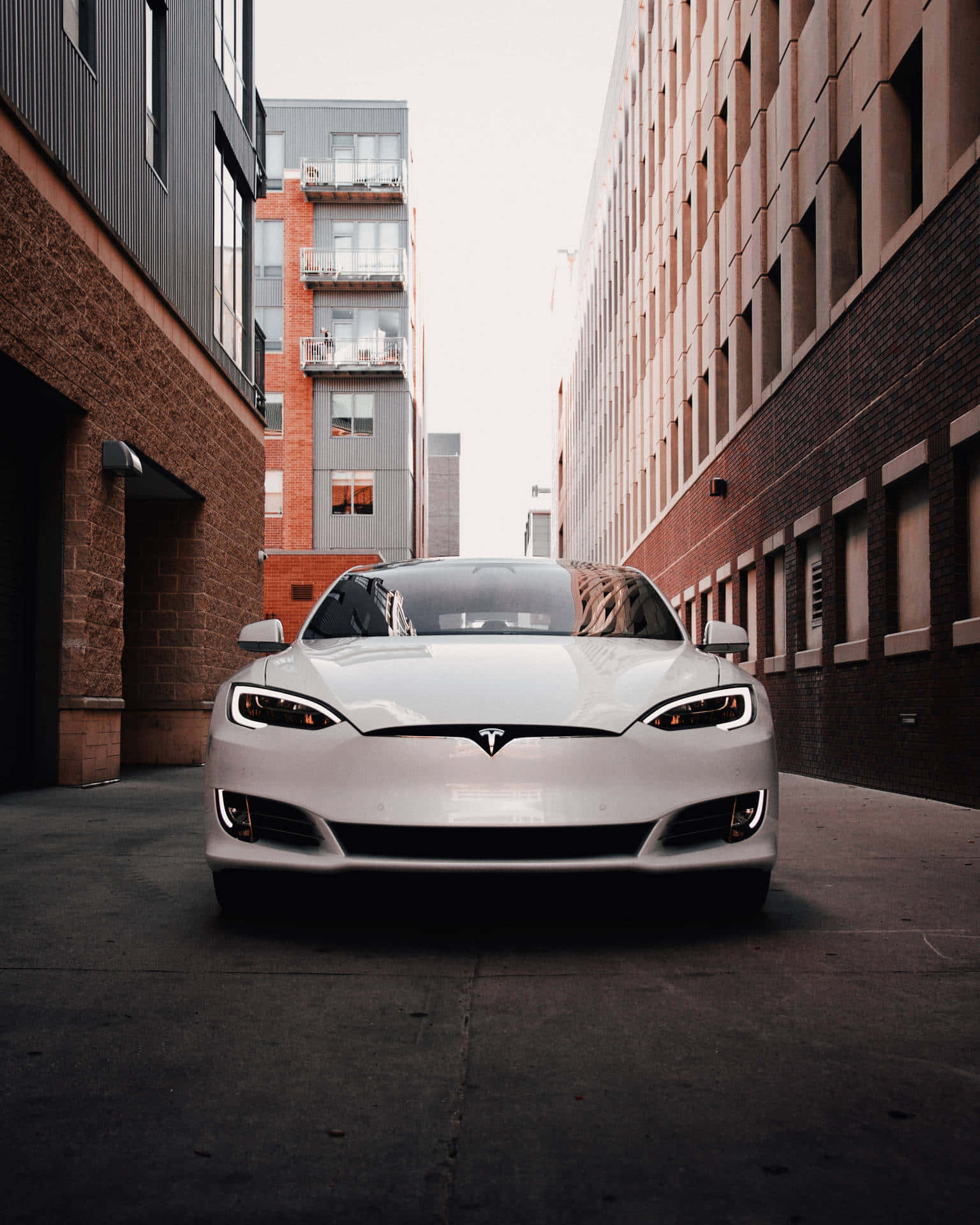 特斯拉 桌布 壁紙 — 一輛時尚的白色Tesla Model S在狹窄的城市小巷中向前，兩旁是磚砌和現代建築，天空多雲。