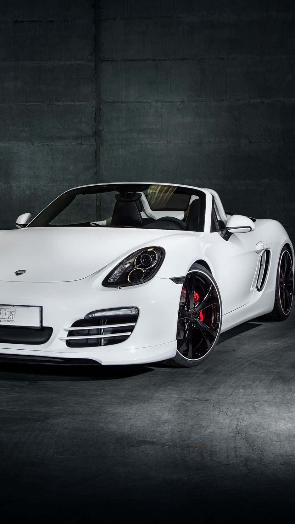 Porsche iPhone 桌布 壁紙 — 一輛時尚的白色保時捷Boxster敞篷跑車，配備黑色輪圈和紅色制動卡鉗，在戲劇性的攝影棚燈光下，停靠在深色混凝土牆前。