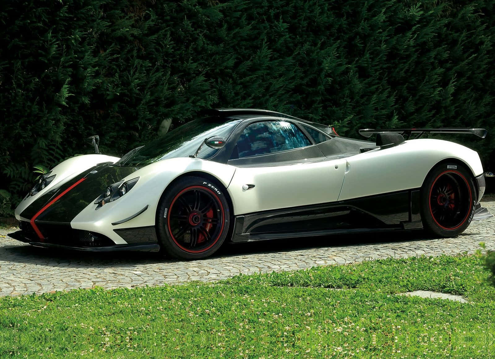 Pagani Zonda Cinque 桌布 壁紙 — 一輛時尚的白色和黑色帕加尼超級跑車，帶有紅色點綴，停在鵝卵石小徑旁，旁邊是綠草和茂密的樹籬。
