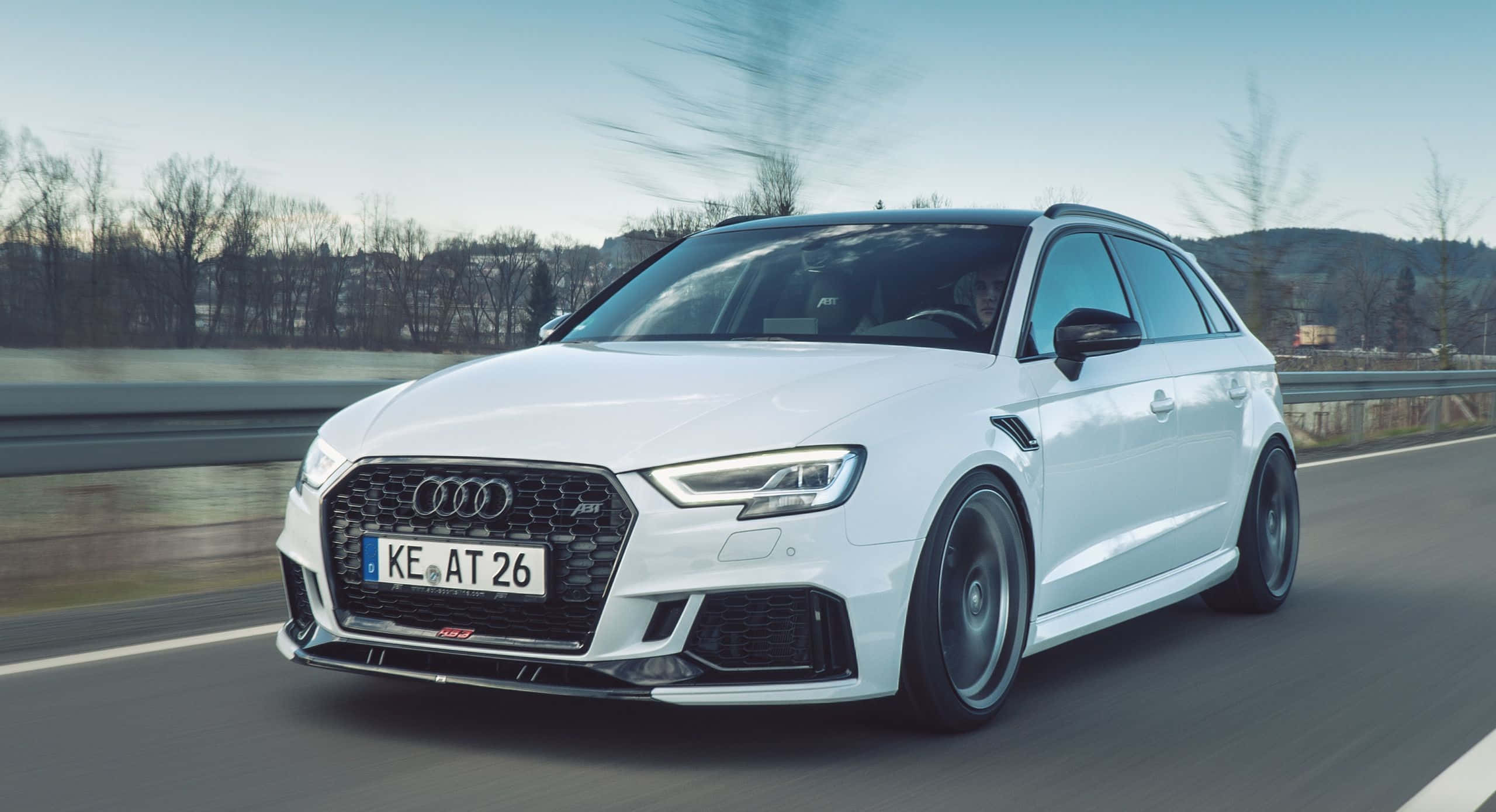 Audi Rs3 桌布 壁紙 — 一輛時尚的白色奧迪RS3 Sportback在高速公路上疾駛，背景是模糊的樹木和山丘，天空晴朗。