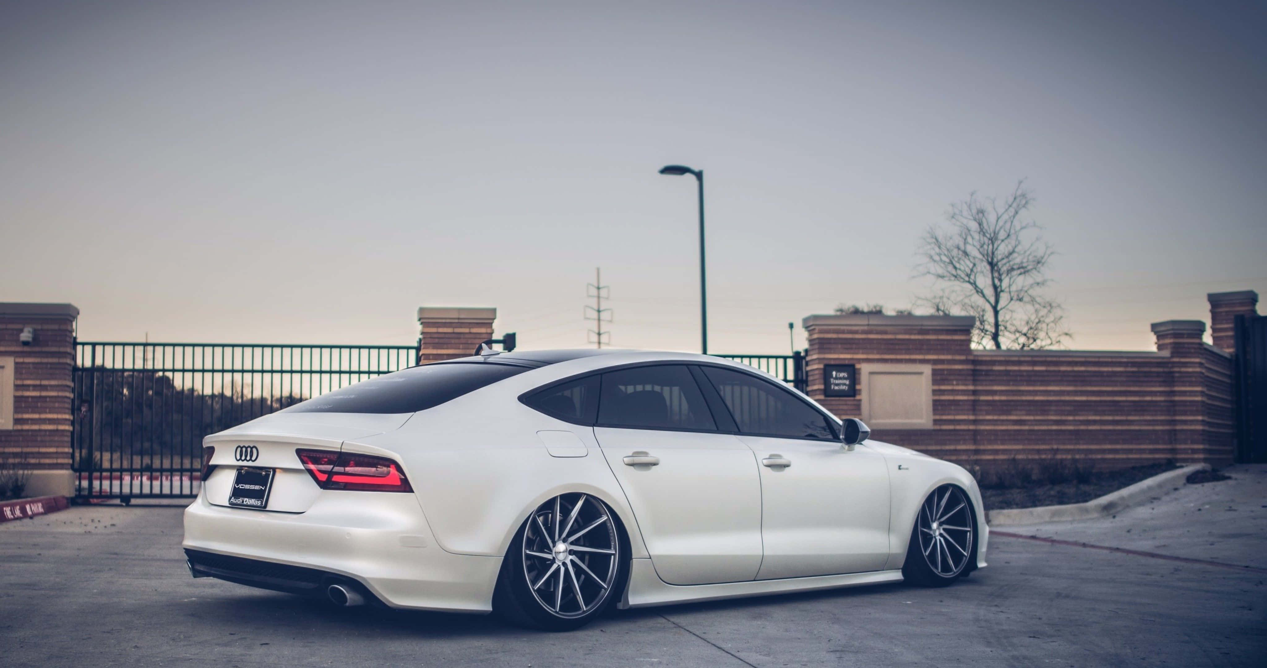 Audi S7 桌布 壁紙 — 一輛時尚的白色奧迪房車配備訂製輪圈，停泊在黃昏時分的混凝土停車場，靠近磚牆和圍欄。