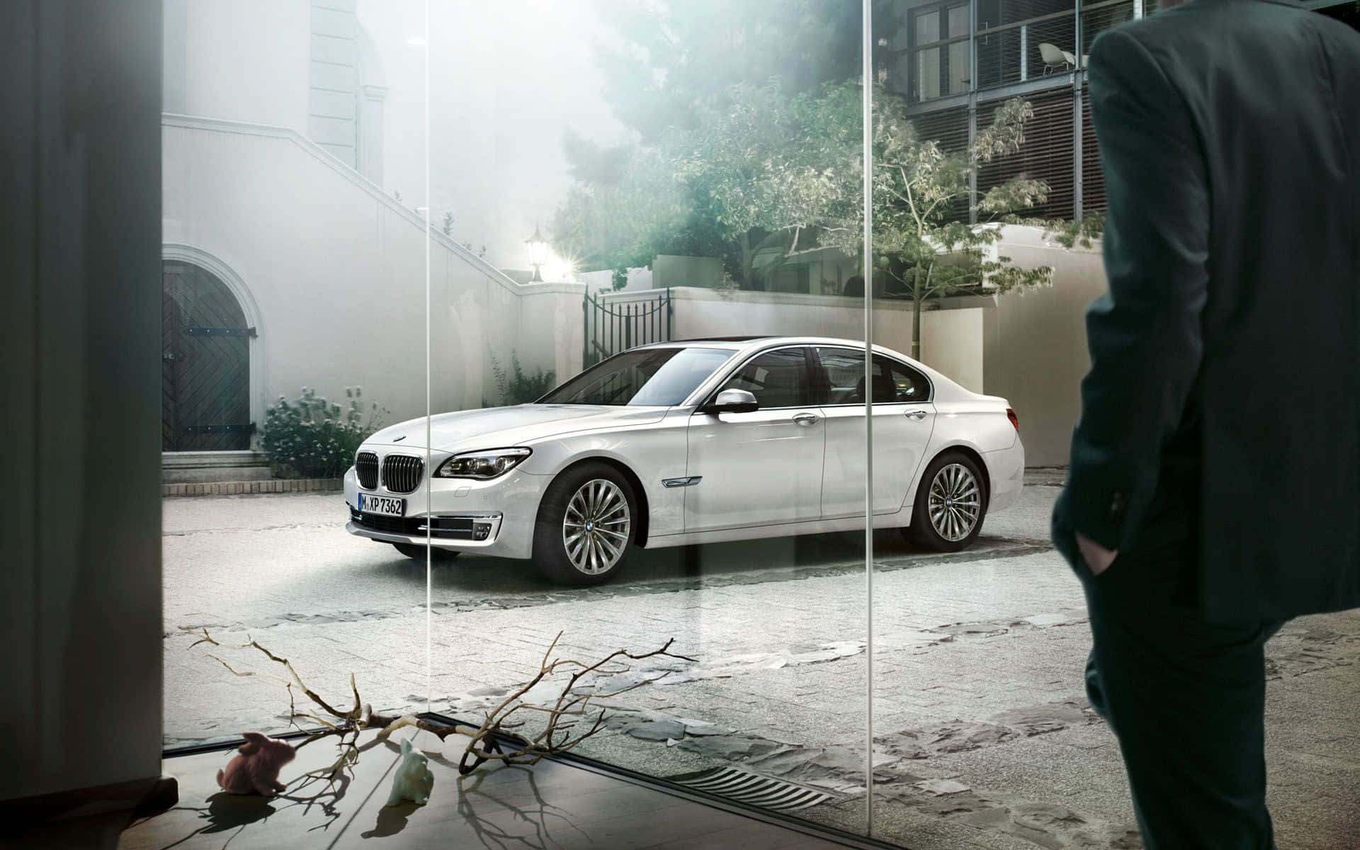 Bmw 7 Series 桌布 壁紙 — 一輛時尚的白色寶馬轎車停在霧氣瀰漫的庭院中，透過破碎的玻璃窗看到，附近有一位身穿西裝的男子站立。