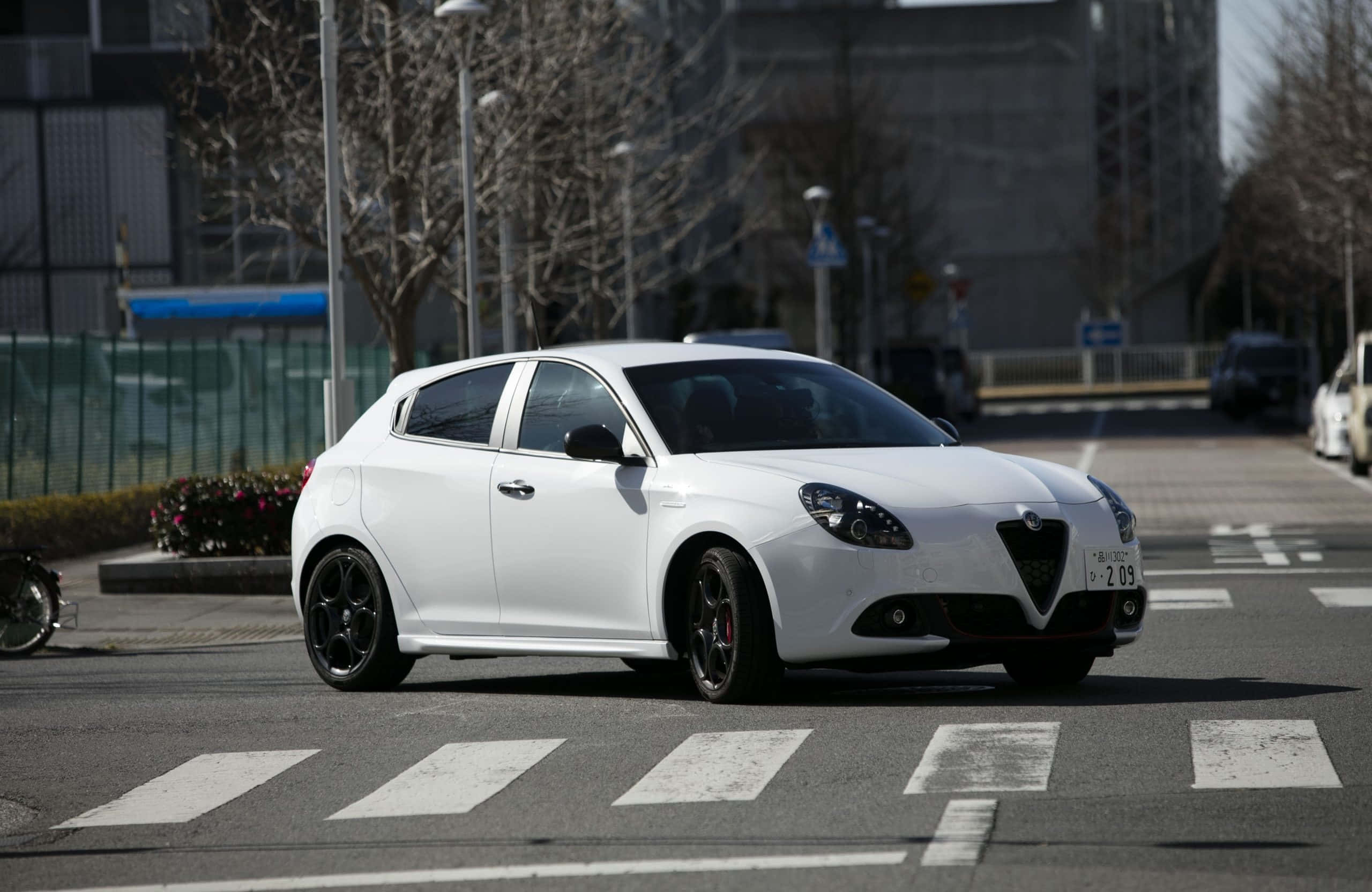 Alfa Romeo Giulietta 桌布 壁紙 — 一輛時尚的白色愛快羅密歐Giulietta配黑色輪圈，在陽光普照的市區街道上行駛，街道上有行人穿越道。