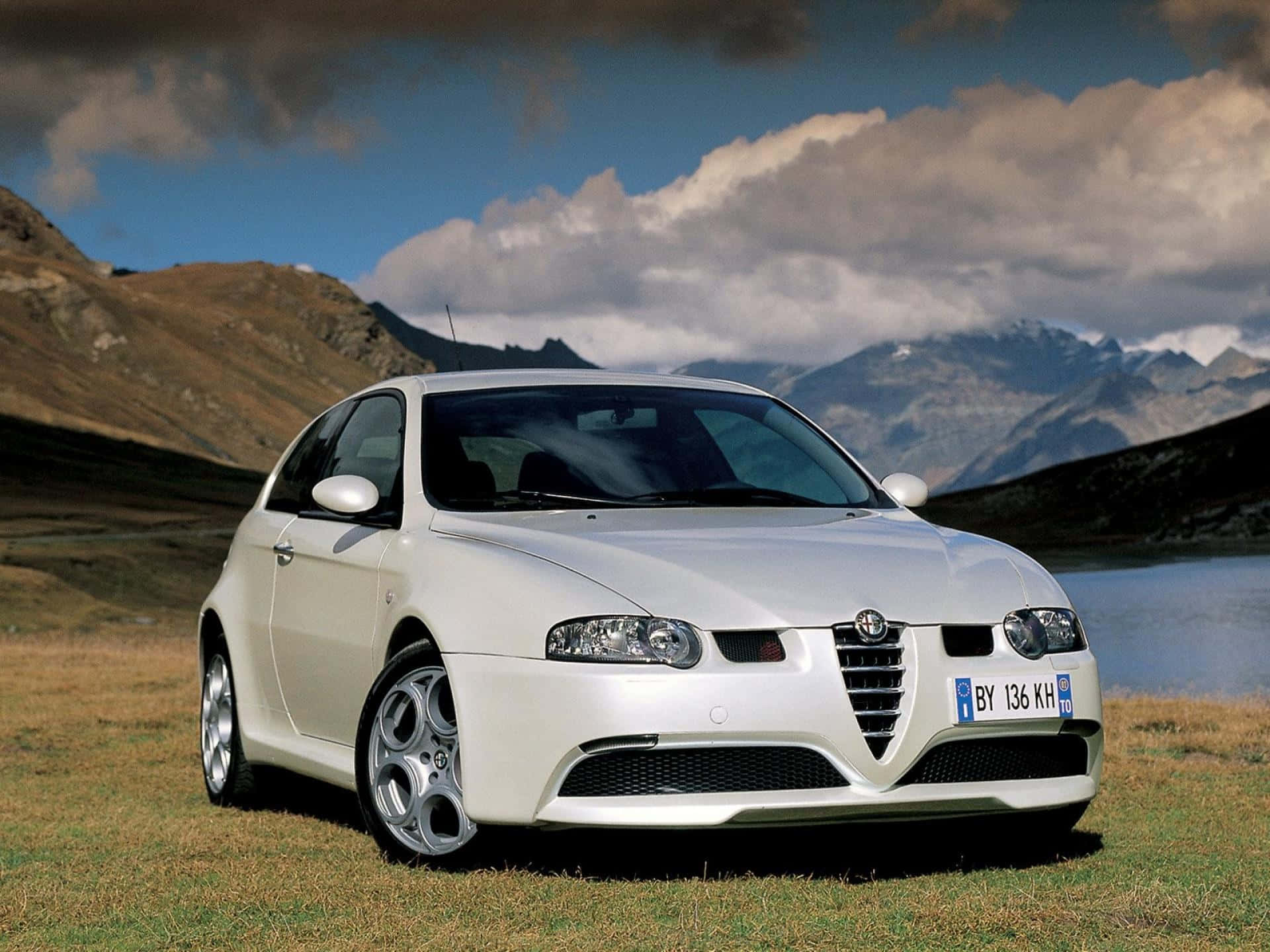 Alfa Romeo 147 桌布 壁紙 — 一輛時尚的白色愛快羅密歐跑車停在草地上，背景是山脈和湖泊，天空多雲。