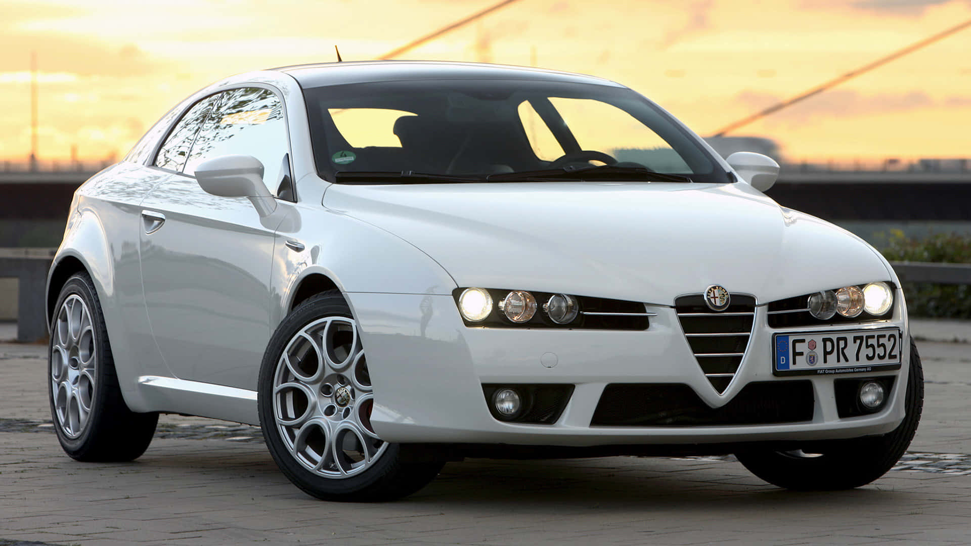 Alfa Romeo Brera 桌布 壁紙 — 一輛時尚的白色愛快羅密歐跑車在金色夕陽下閃耀於鋪砌路面上。
