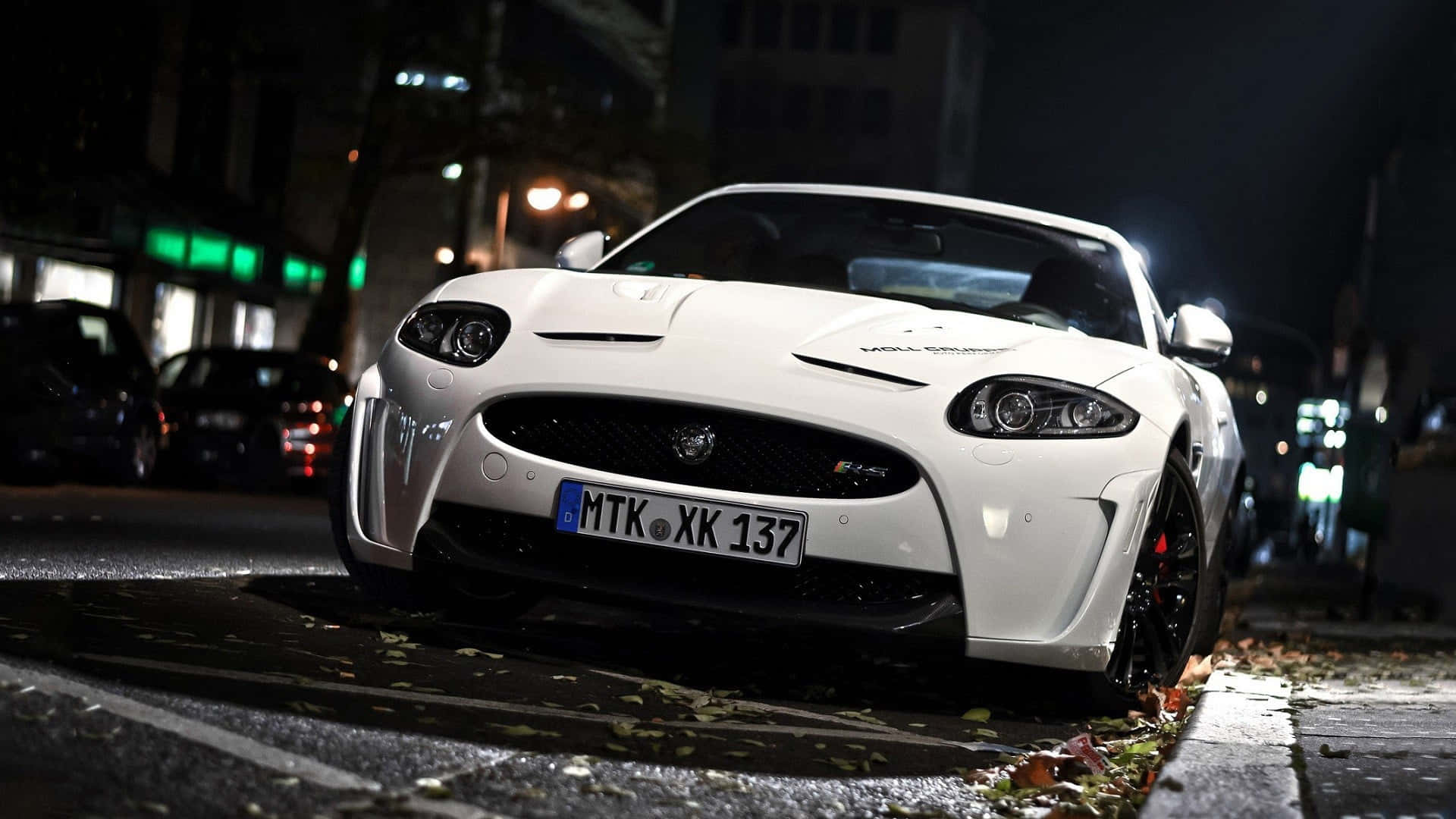 Jaguar Xkr 桌布 壁紙 — 一輛時尚的白色捷豹跑車停泊在濕漉漉的城市街道上，夜晚中車頭燈閃爍，周圍散落著秋葉。