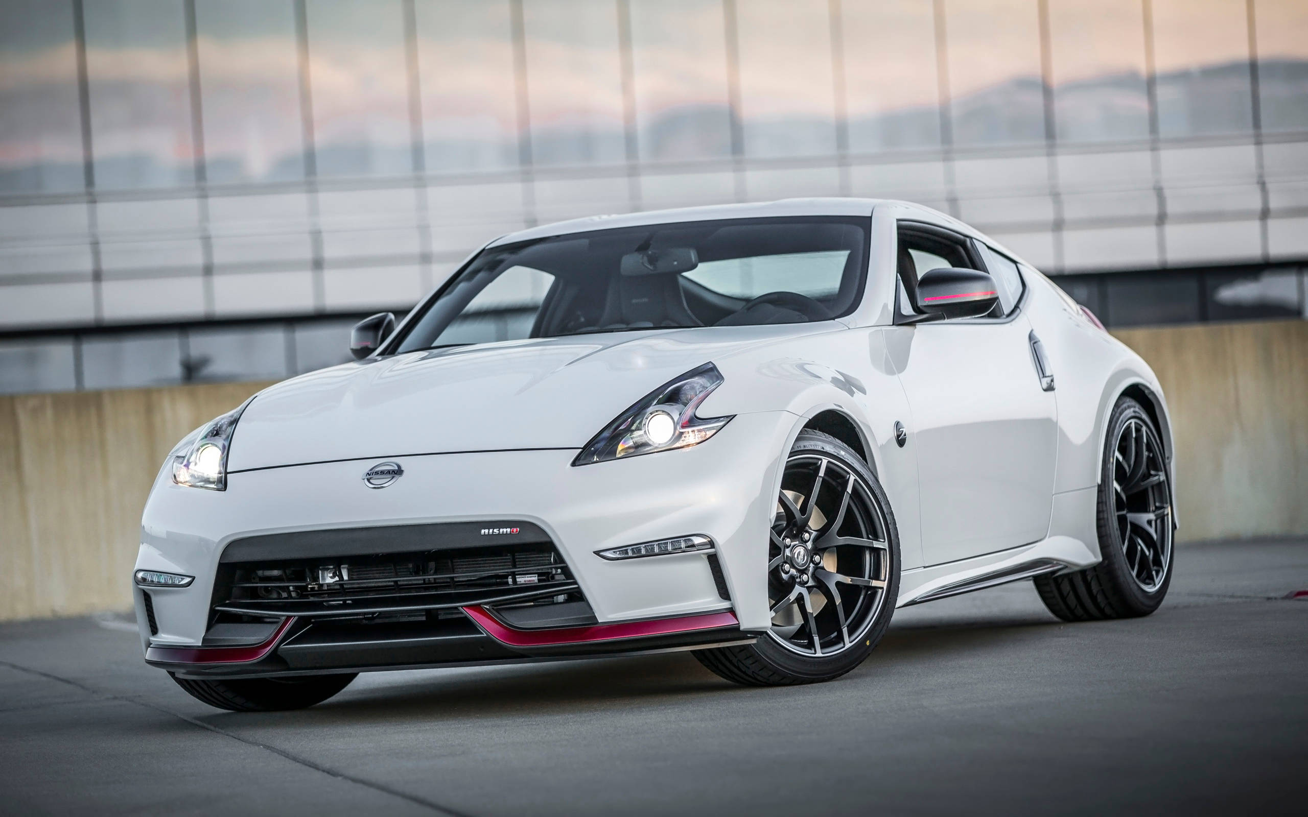 Nissan 370z 桌布 壁紙 — 一輛時尚的白色日產370Z跑車，配備黑色輪圈和紅色點綴，停放在黃昏時分現代玻璃建築前的混凝土表面上。