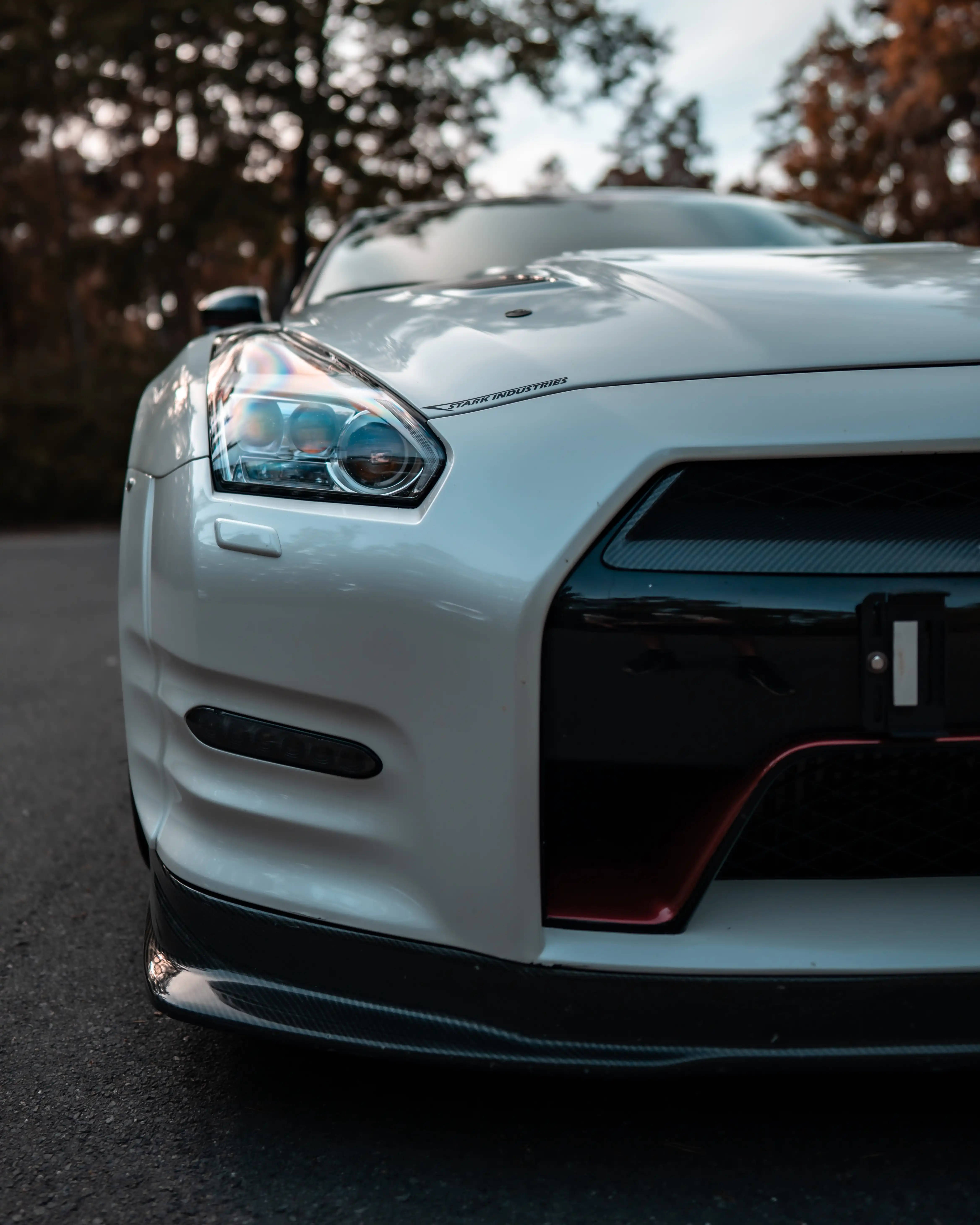 Nissan 桌布 壁紙 — 一輛時尚的白色日產GT-R跑車，帶有碳纖維飾邊和紅色飾條，停在秋天的樹下的瀝青路上。