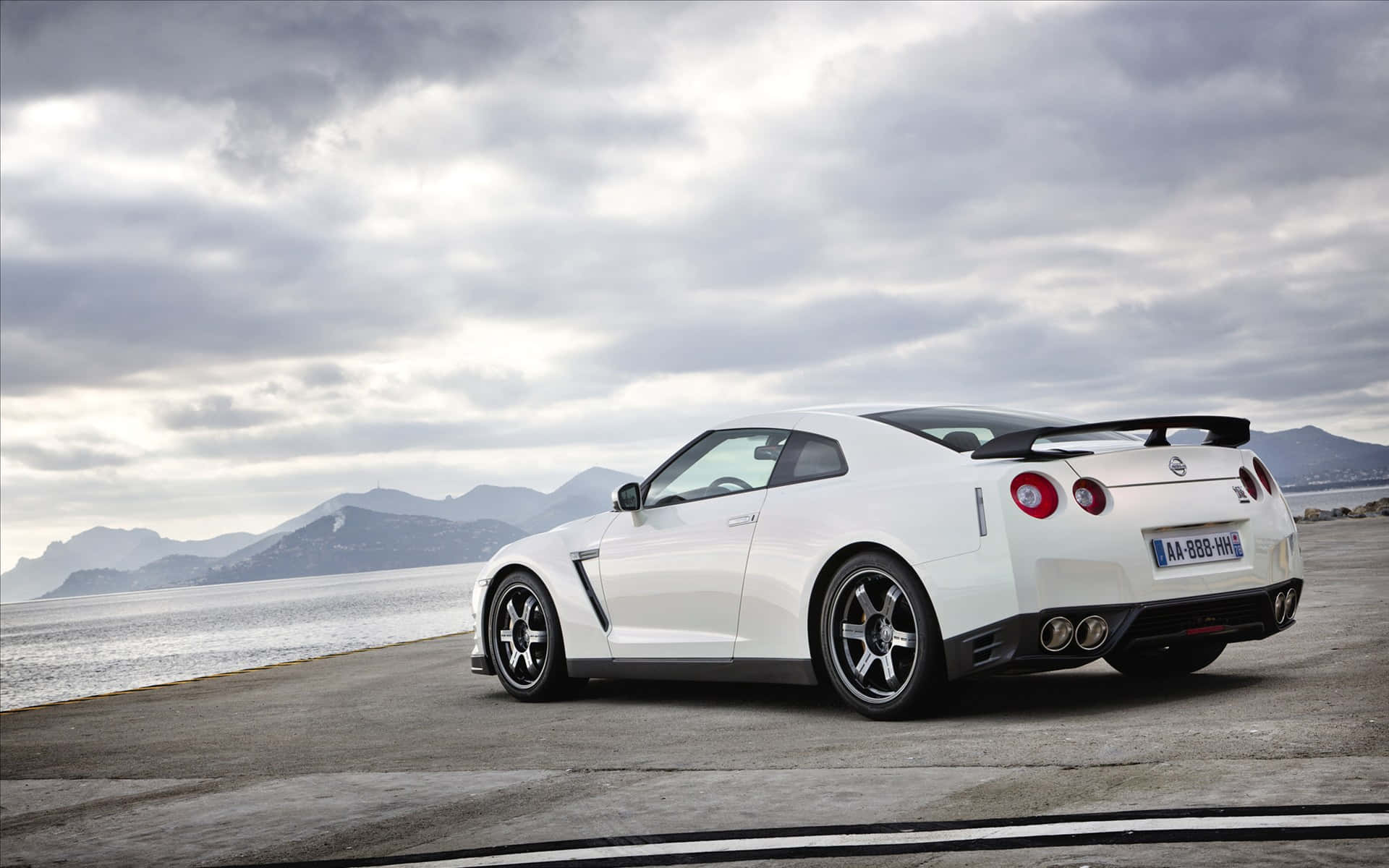 Gtr 桌布 壁紙 — 一輛時尚的白色日產GT-R跑車停泊在沿海公路上，背景是山脈和陰天。