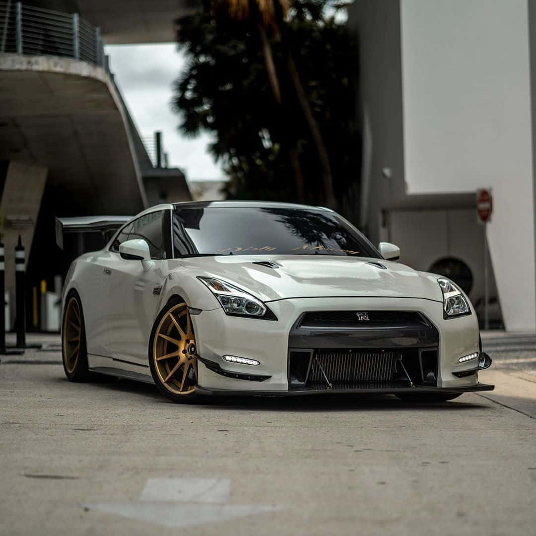 日產 桌布 壁紙 — 一輛時尚的白色日產GT-R配金色輪圈，停泊在現代混凝土天橋下，背景有棕櫚樹。