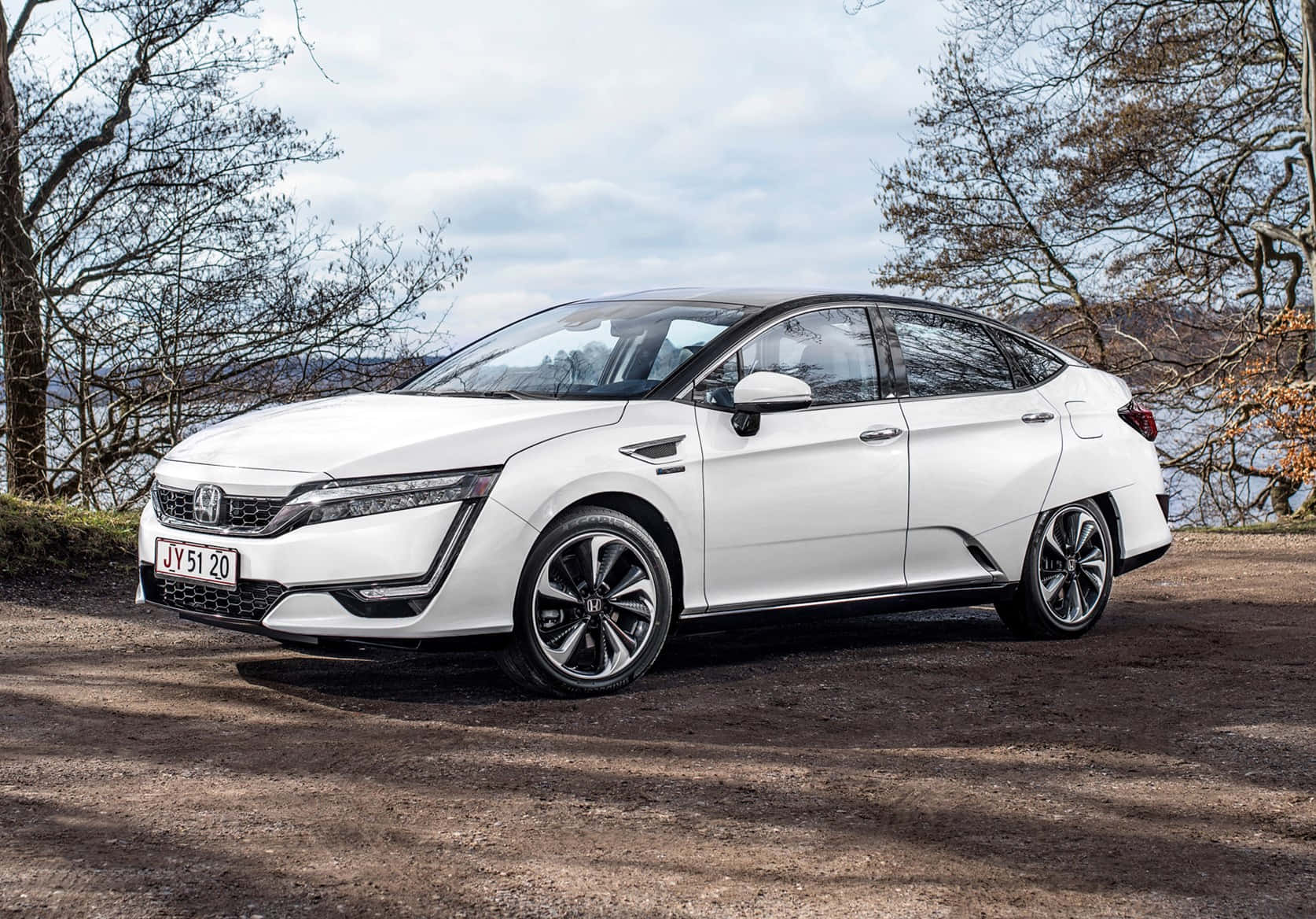 Honda Clarity 桌布 壁紙 — 一輛時尚的白色本田Clarity插電式混合動力車停泊在湖邊的泥路上，周圍是光禿禿的樹木，天空多雲。