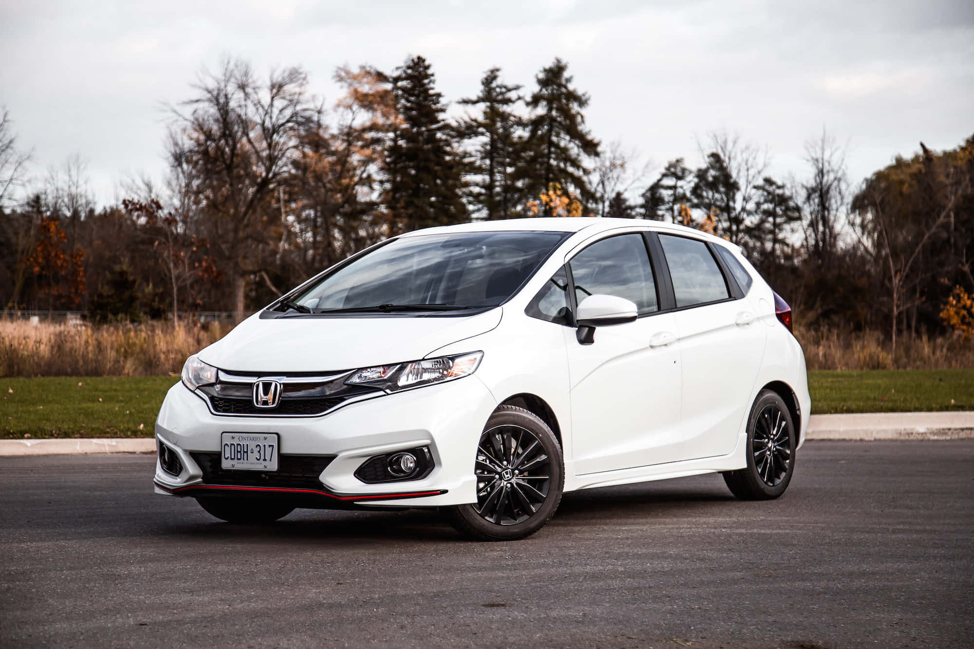 Honda Fit 桌布 壁紙 — 一輛時尚的白色本田Fit，配黑色輪圈，停泊在瀝青路上，旁邊是秋天的樹木，天空多雲。