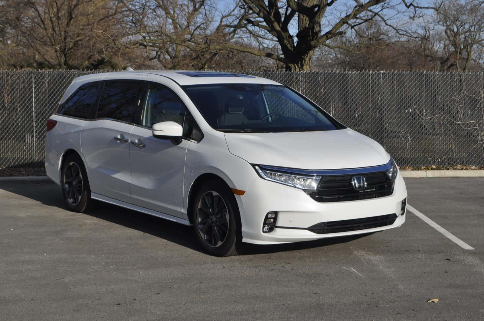 Honda Odyssey 桌布 壁紙 — 一輛時尚的白色本田Odyssey小型貨車停放在陽光明媚的瀝青停車場，背景有光禿禿的樹木和鐵絲網圍欄。
