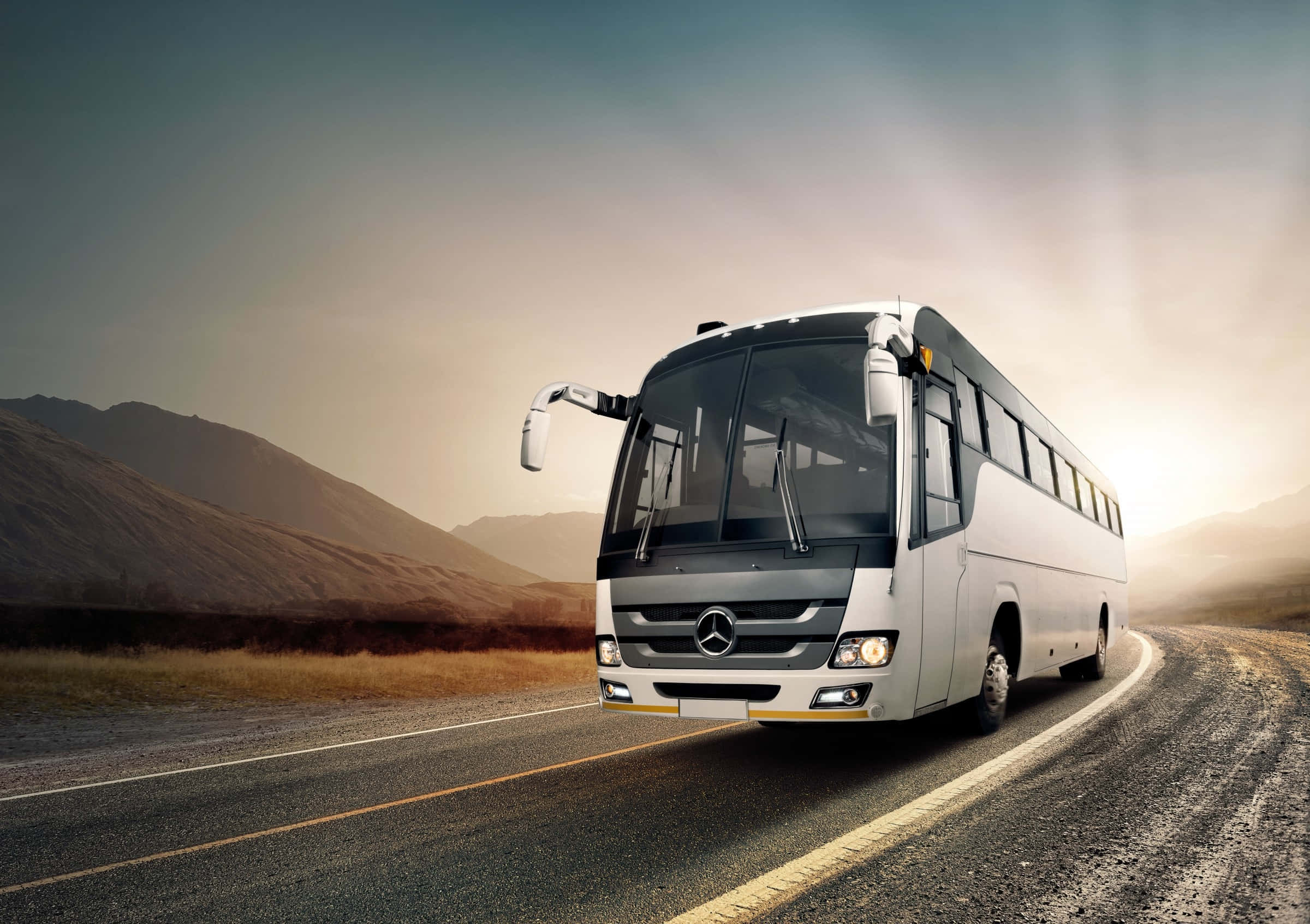 Bus 桌布 壁紙 — 一輛時尚的白色梅賽德斯巴士沿著蜿蜒的道路行駛，穿越乾燥的山脈，天空籠罩著朦朧的金色。