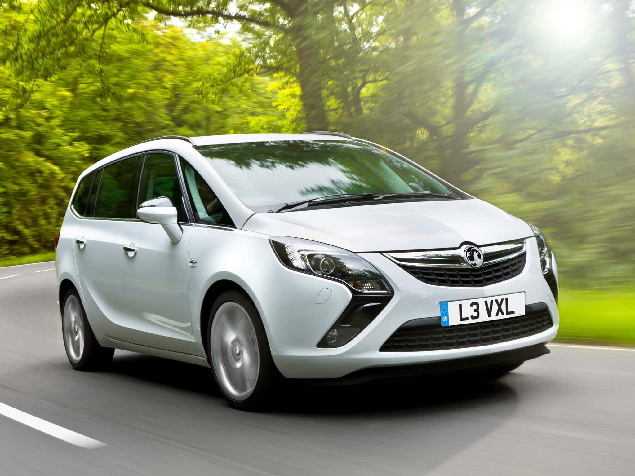 Opel Zafira 桌布 壁紙 — 一輛時尚的白色歐寶Zafira在陽光明媚的森林道路上快速行駛，模糊的綠色樹葉從旁邊掠過。