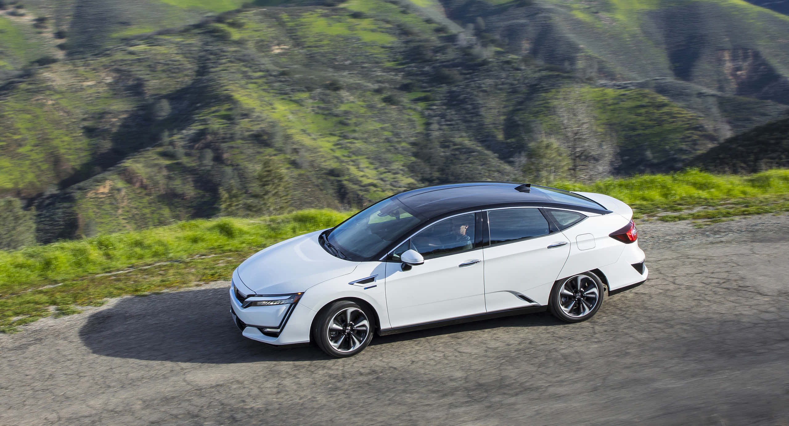 Honda Clarity 桌布 壁紙 — 一輛時尚的白色混合動力車，黑色車頂，沿著蜿蜒的山路行駛，周圍是鬱鬱蔥蔥的綠色山丘。