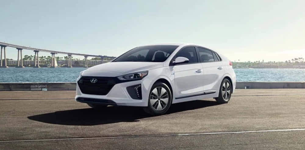Compact Car 桌布 壁紙 — 一輛時尚的白色現代 Ioniq 混合動力車停在海濱道路上，背景是橋樑和寧靜的藍色海水，在明亮的日光下。