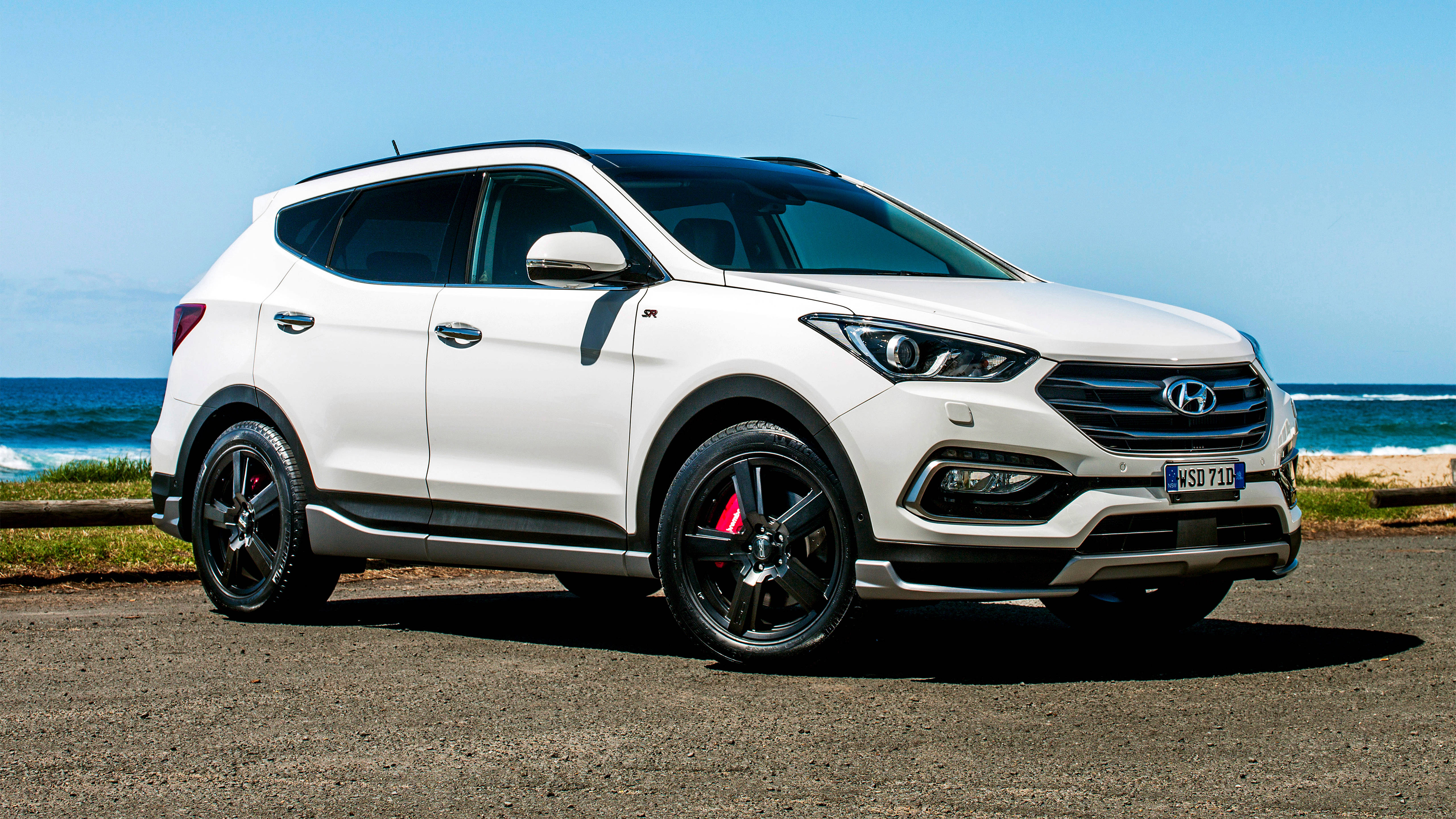 Hyundai 桌布 壁紙 — 一輛時尚的白色現代Santa Fe SUV，配備黑色輪圈和紅色制動卡鉗，停泊在海洋旁邊的碎石路上，天空清澈湛藍。