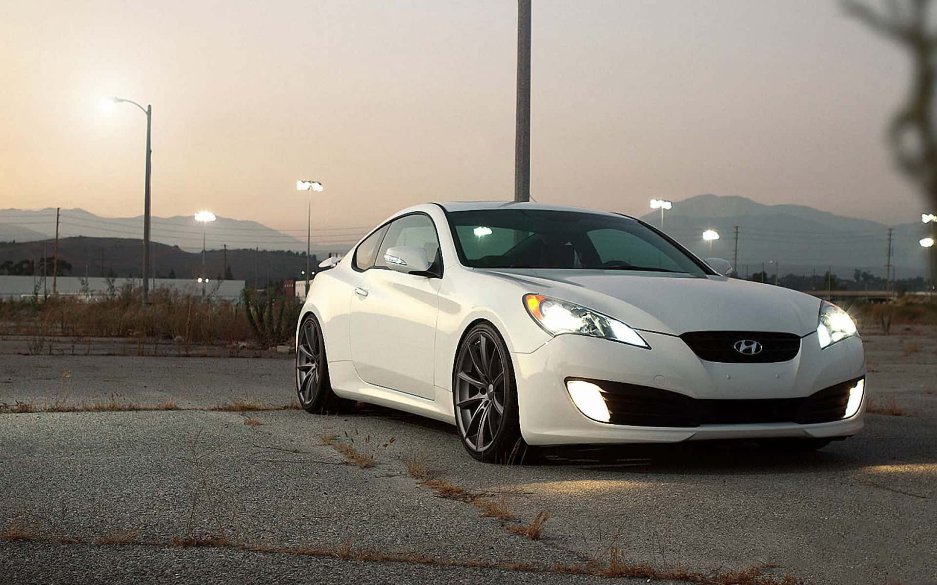Hyundai Genesis 桌布 壁紙 — 一輛時尚的白色現代轎跑車，車頭燈發亮，停在黃昏時分龜裂的柏油路上，遠方有山脈和路燈。