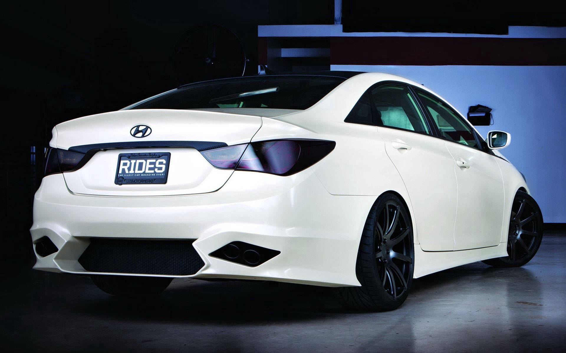 Hyundai 桌布 壁紙 — 一輛時尚的白色現代轎車，配備黑色輪圈和定制排氣管，在昏暗的車庫中，在戲劇性的藍色燈光下展示。