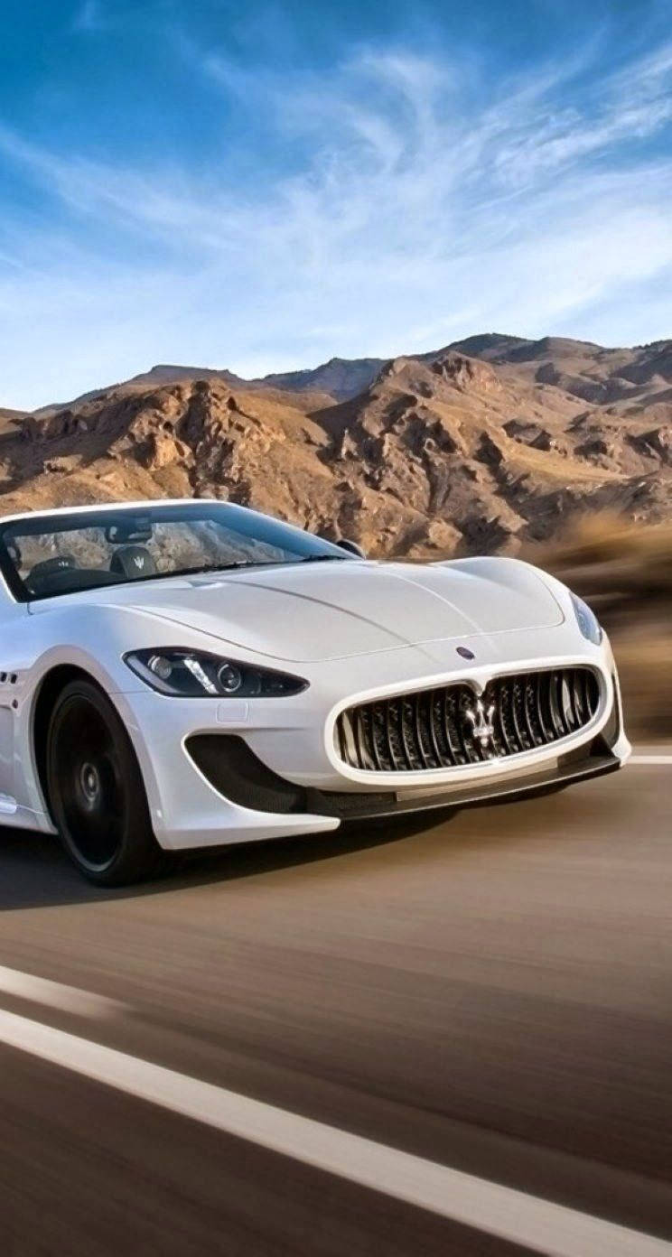 汽車 iPhone 壁紙 桌布 壁紙 — 一輛時尚的白色瑪莎拉蒂跑車在藍天白雲下，沿著崎嶇山脈環繞的沙漠公路飛馳。