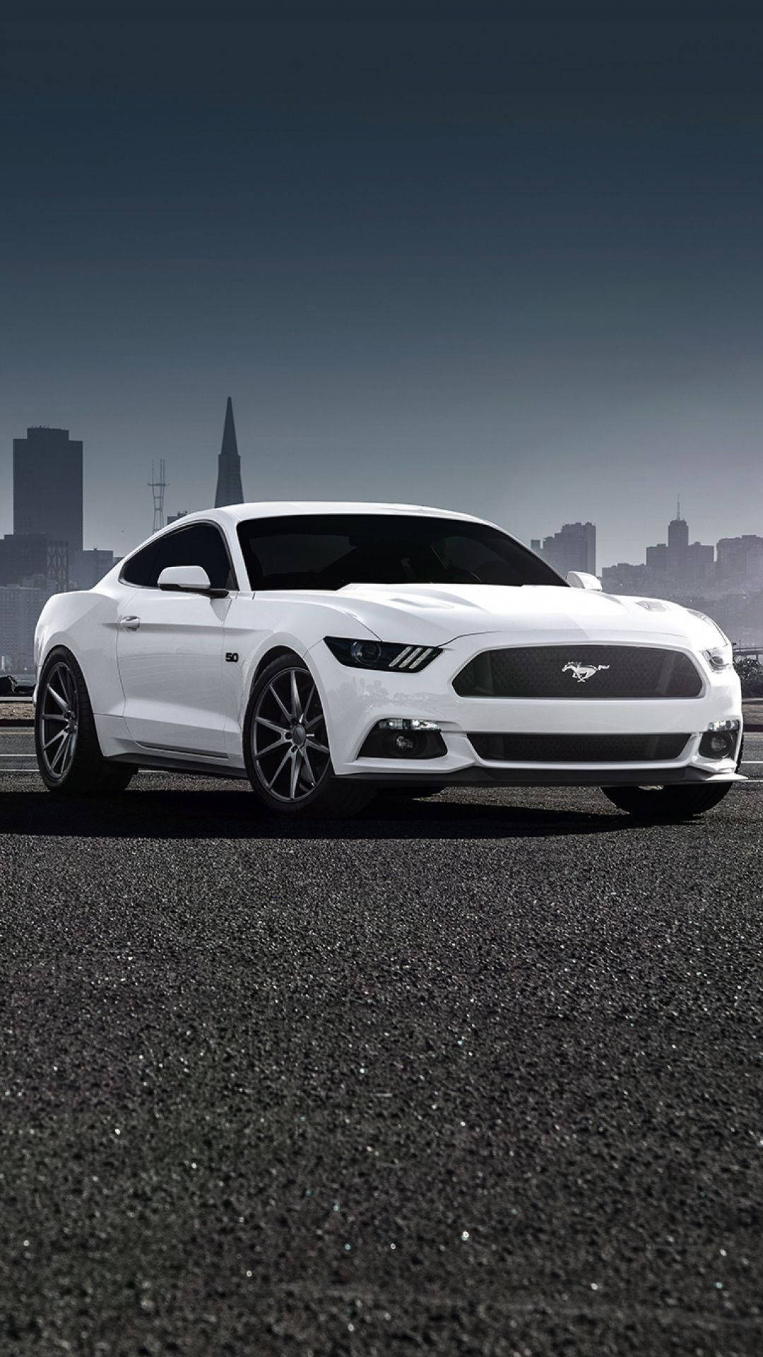 ford iPhone 桌布 壁紙 — 一輛時尚的白色福特野馬跑車停在柏油路上，背景是朦朧的城市天際線。