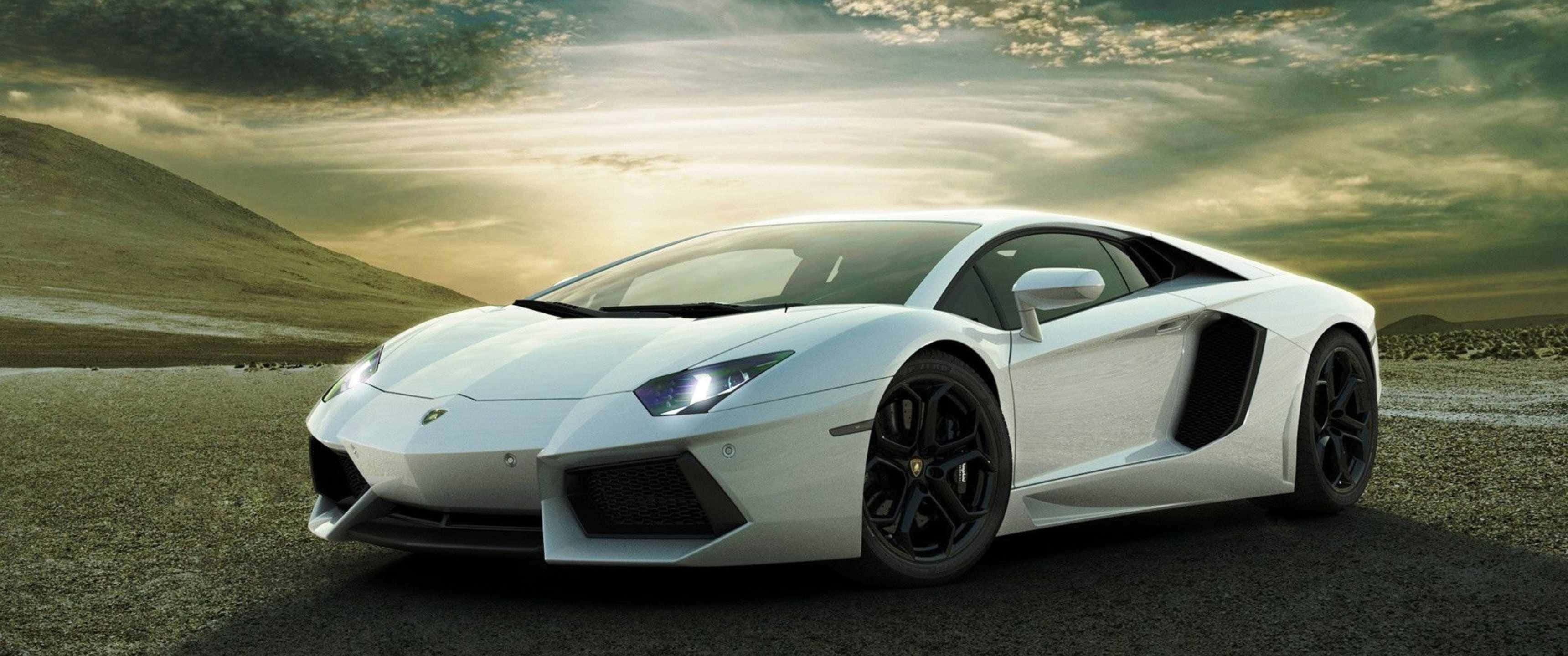 3440x1440p Lamborghini 桌布 壁紙 — 一輛時尚的白色藍寶堅尼 Aventador 停泊在碎石路上，背景是戲劇性的金色日落天空。