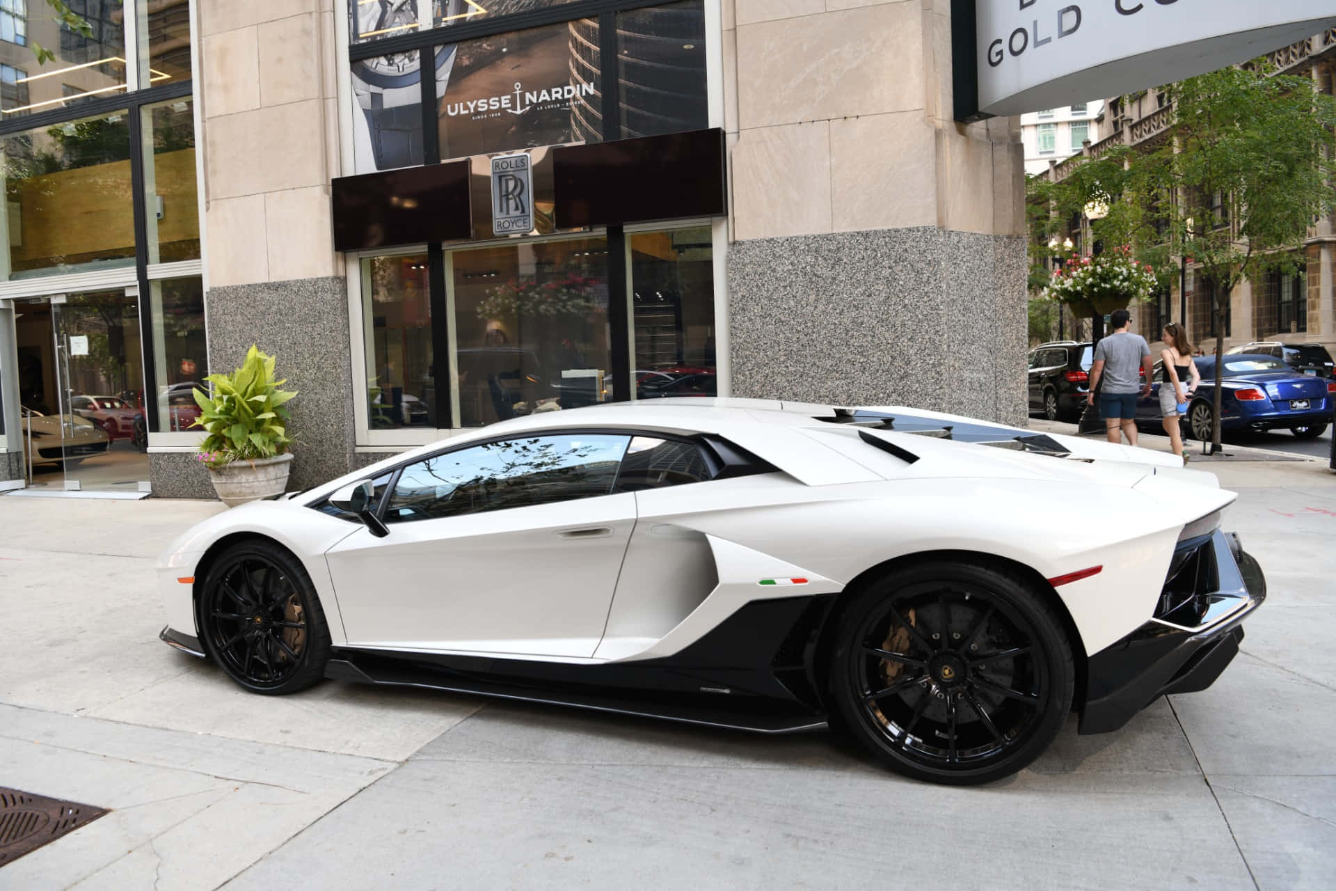 跑車 桌布 壁紙 — 一輛時尚的白色藍寶堅尼Aventador，帶有黑色飾邊，停泊在豪華商店附近的城市人行道上。
