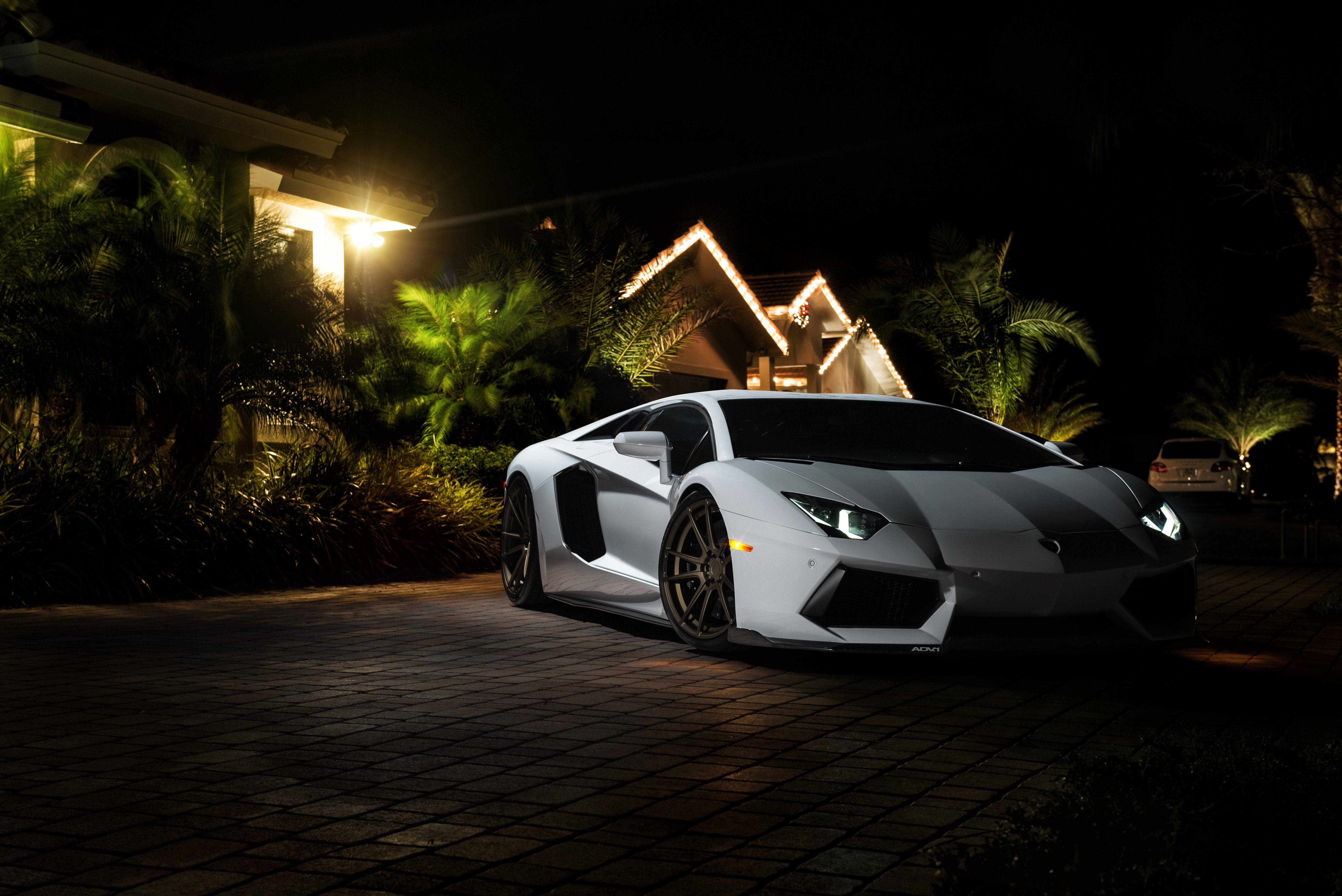 高清 桌布 壁紙 — 一輛時尚的白色藍寶堅尼Aventador在夜間停泊在磚砌車道上，被溫暖的房屋燈光和閃爍的車頭燈照亮，背景是黑暗的熱帶植物。