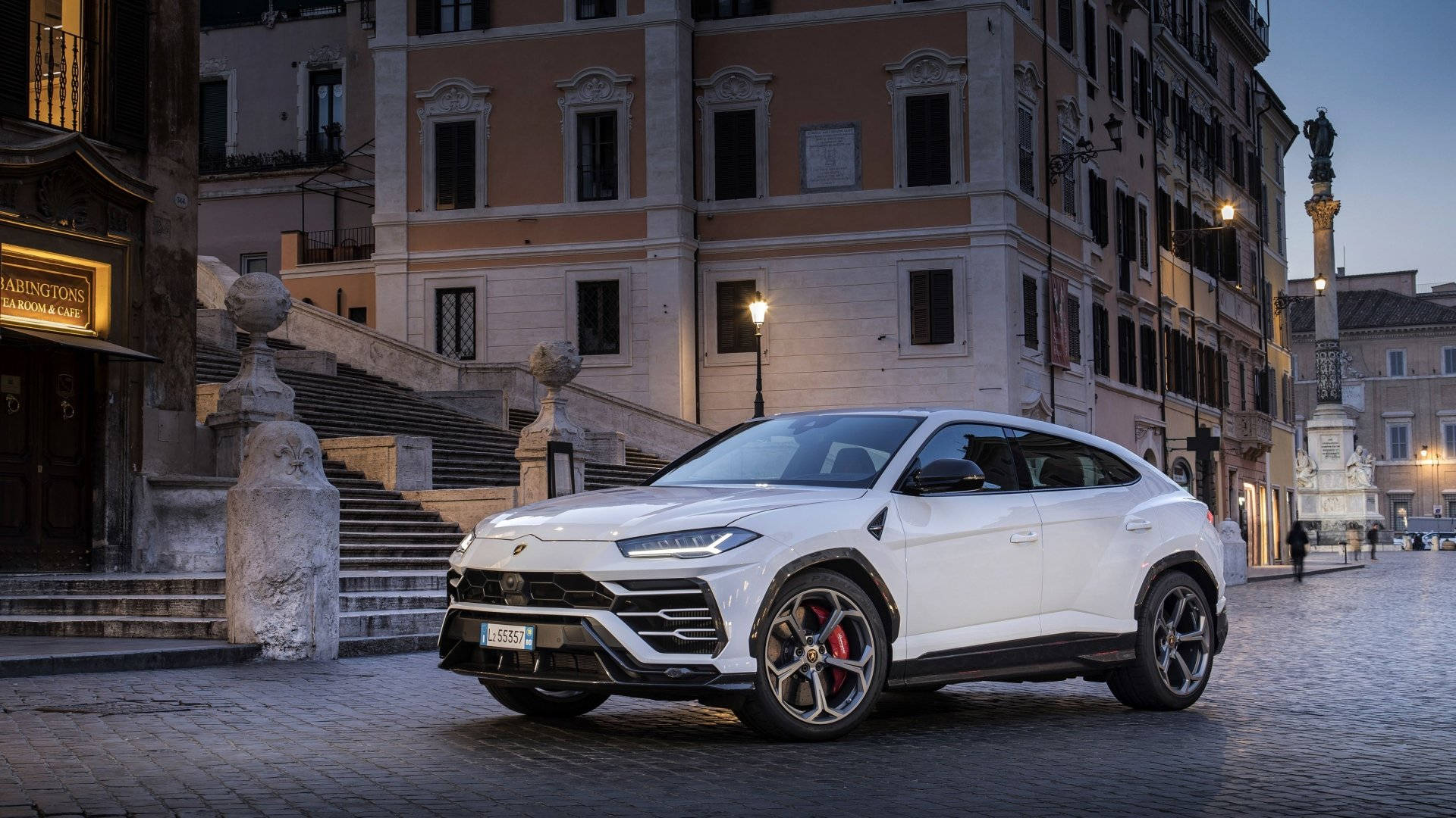 lambo 桌布 壁紙 — 一輛時尚的白色藍寶堅尼Urus SUV停泊在黃昏時分的鵝卵石街道上，背景是歷史建築和溫暖的路燈。