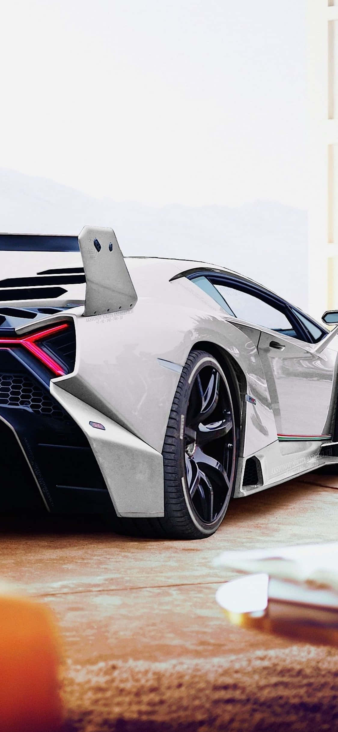 iPhone X Lamborghini 桌布 壁紙 — 一輛時尚的白色藍寶堅尼跑車，帶有發光的紅色尾燈，停放在室內靠近可欣賞山景的窗戶旁。