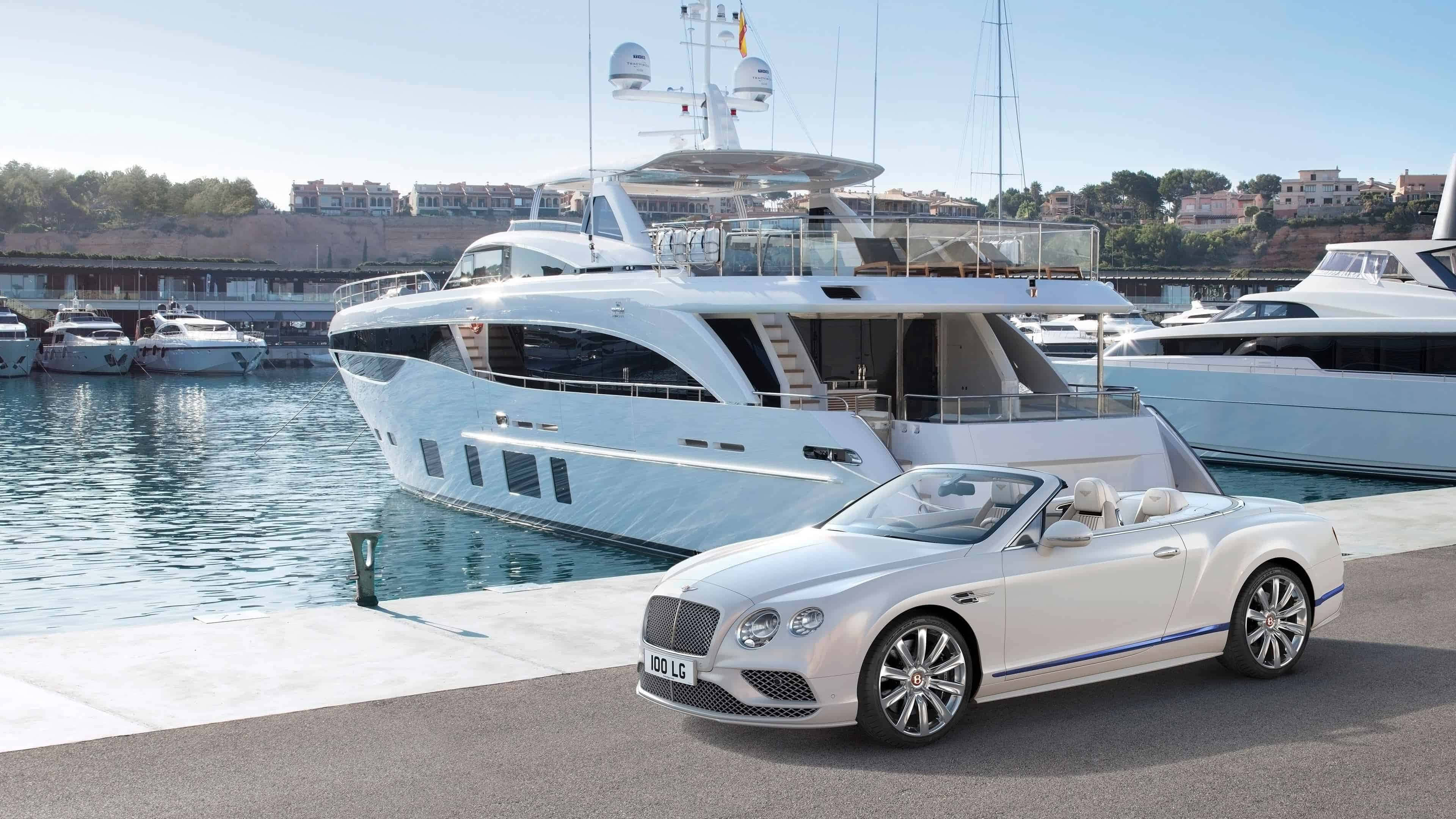 Bentley Continental Gt Convertible 桌布 壁紙 — 一輛時尚的白色賓利敞篷車停泊在豪華遊艇旁邊，位於陽光普照的碼頭，清澈的藍色海水和遠處的建築物。