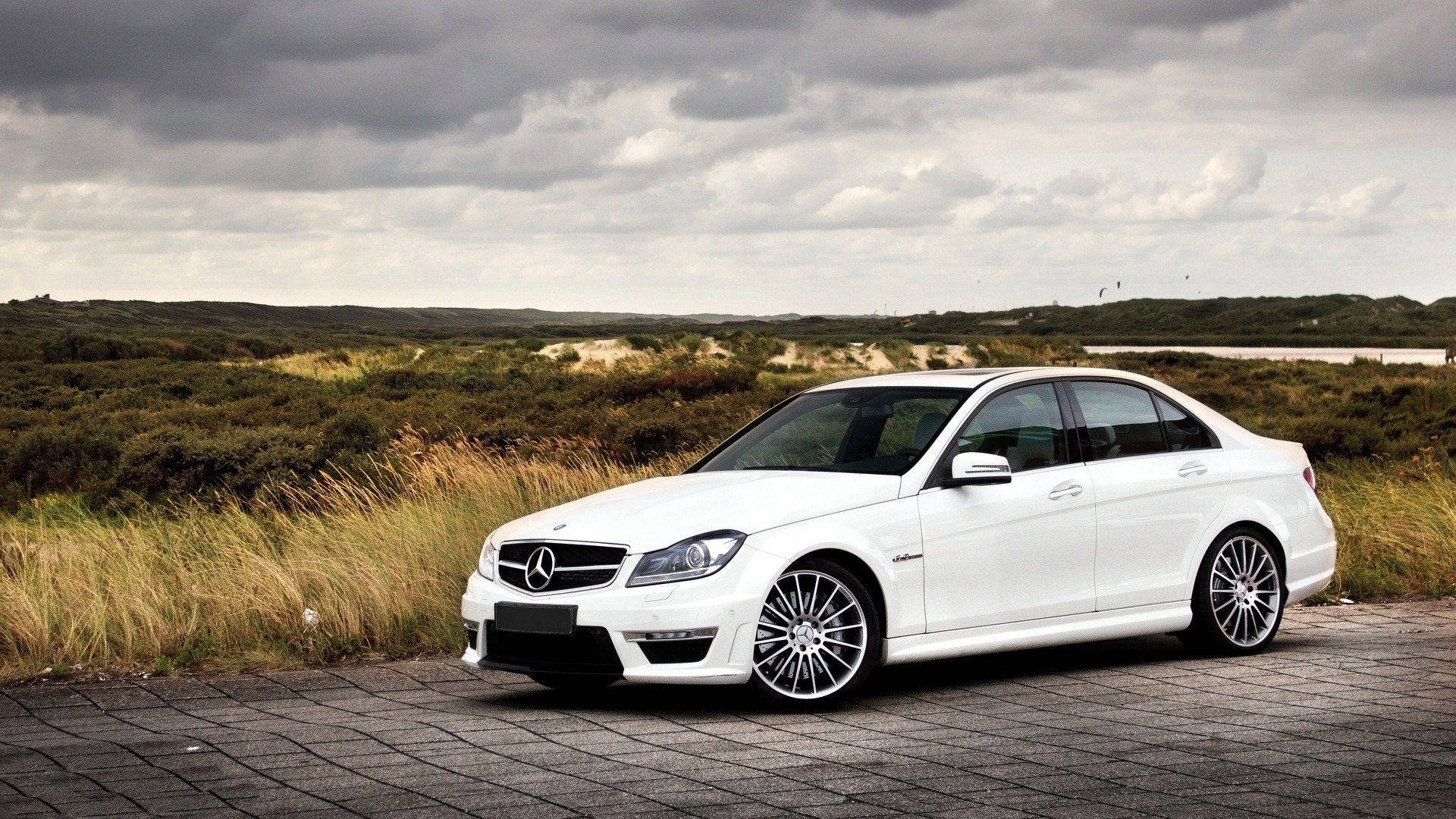 Mercedes 桌布 壁紙 — 一輛時尚的白色賓士轎車停放在鋪砌小徑上，旁邊是高草，天空多雲。