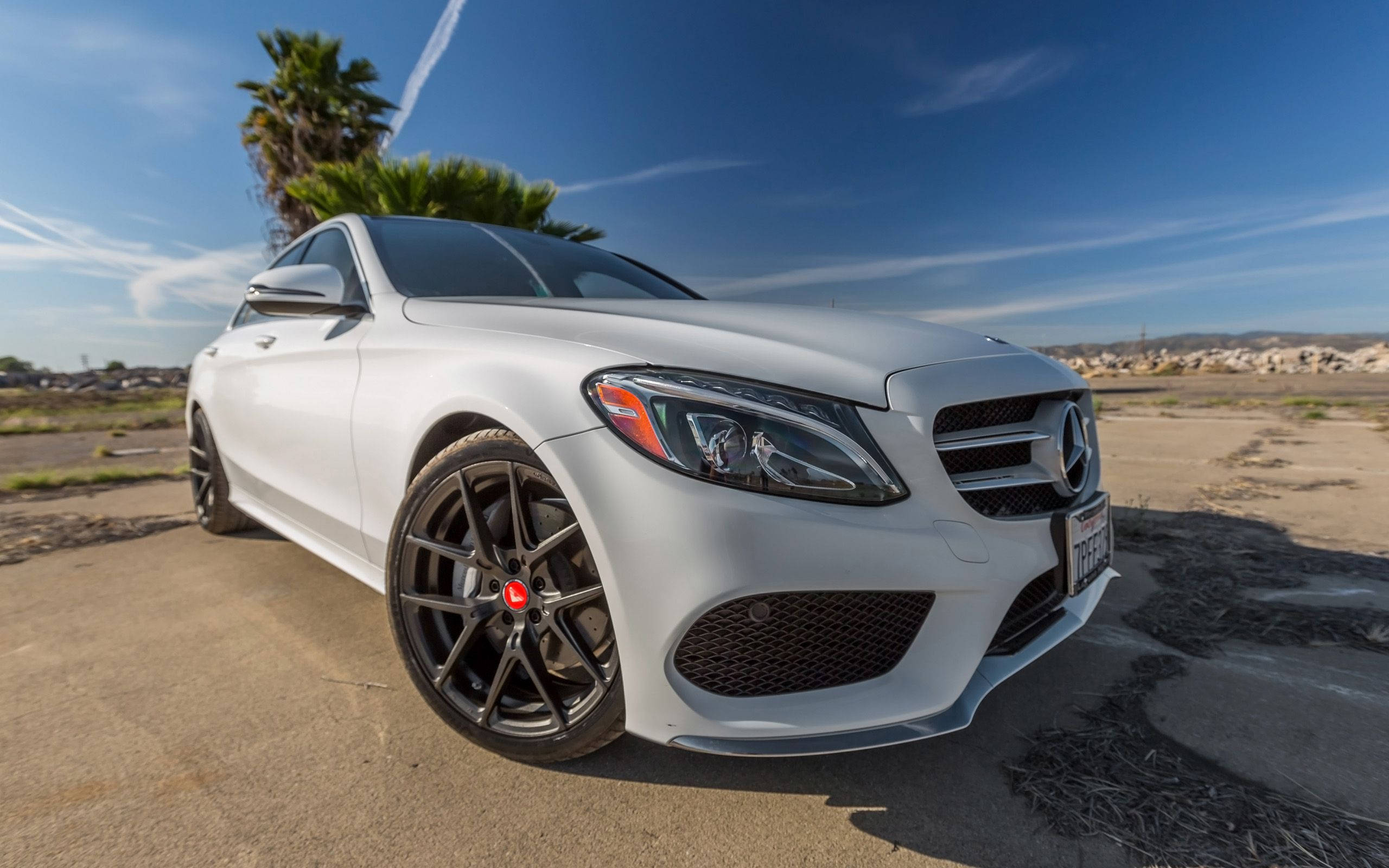 Mercedes Benz C300 桌布 壁紙 — 一輛時尚的白色賓士運動轎車，配備黑色輪圈，停放在陽光普照的混凝土停車場上，背景是藍天白雲和棕櫚樹。