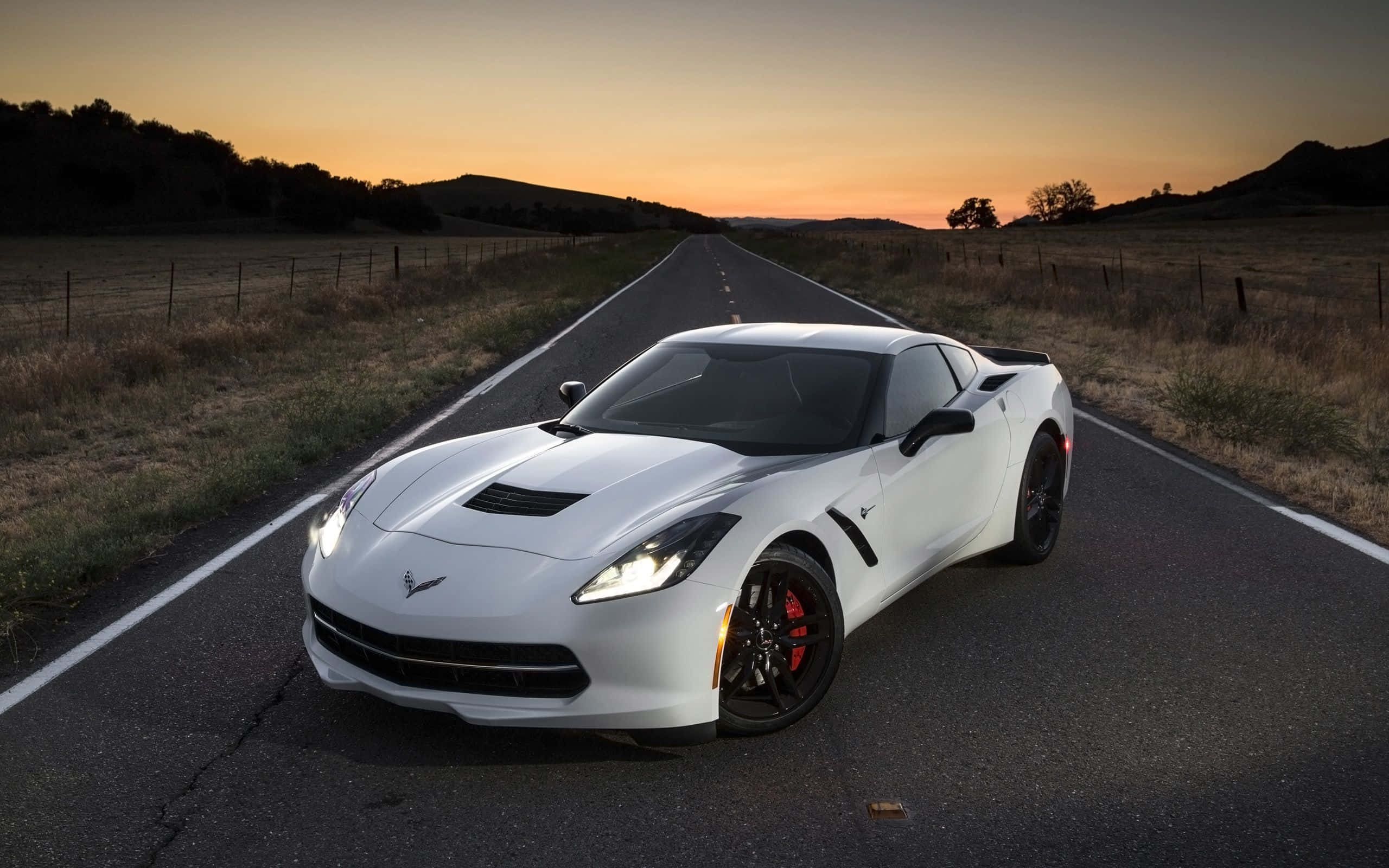 Chevrolet Corvette Stingray 桌布 壁紙 — 一輛時尚的白色跑車，車頭燈閃爍，在日落時分停在一條漫長的鄉村道路上。