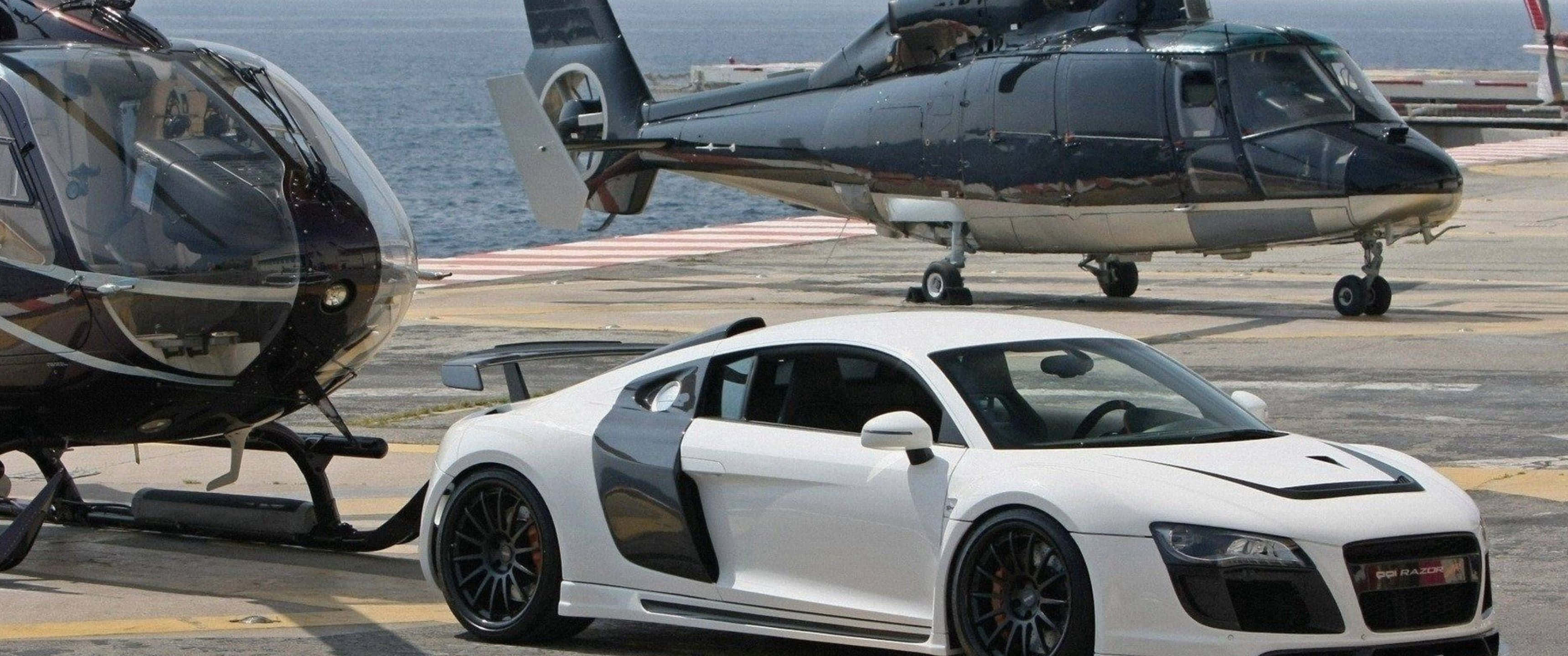 3440x1440p Helicopters 桌布 壁紙 — 一輛時尚的白色跑車停放在兩架黑色直升機旁邊，位於海邊停機坪，在明亮的日光下。