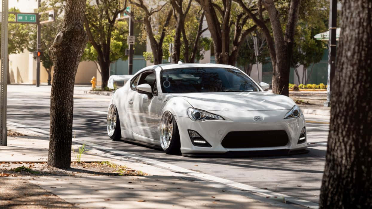 白色跑車停放在陽光明媚的城市街道上，樹木投下陰影。 — Toyota Supra Mk4 壁紙