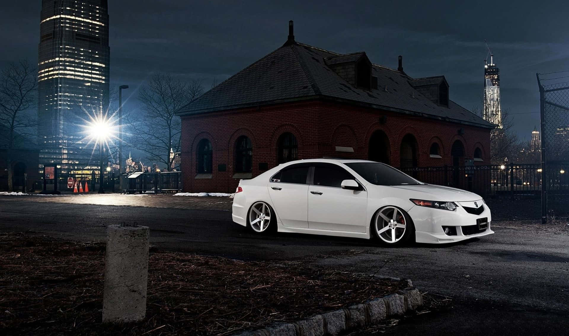 Acura Tsx 桌布 壁紙 — 一輛時尚的白色跑車在夜間停在一棟磚砌建築旁，背景中城市摩天大樓閃爍著光芒。