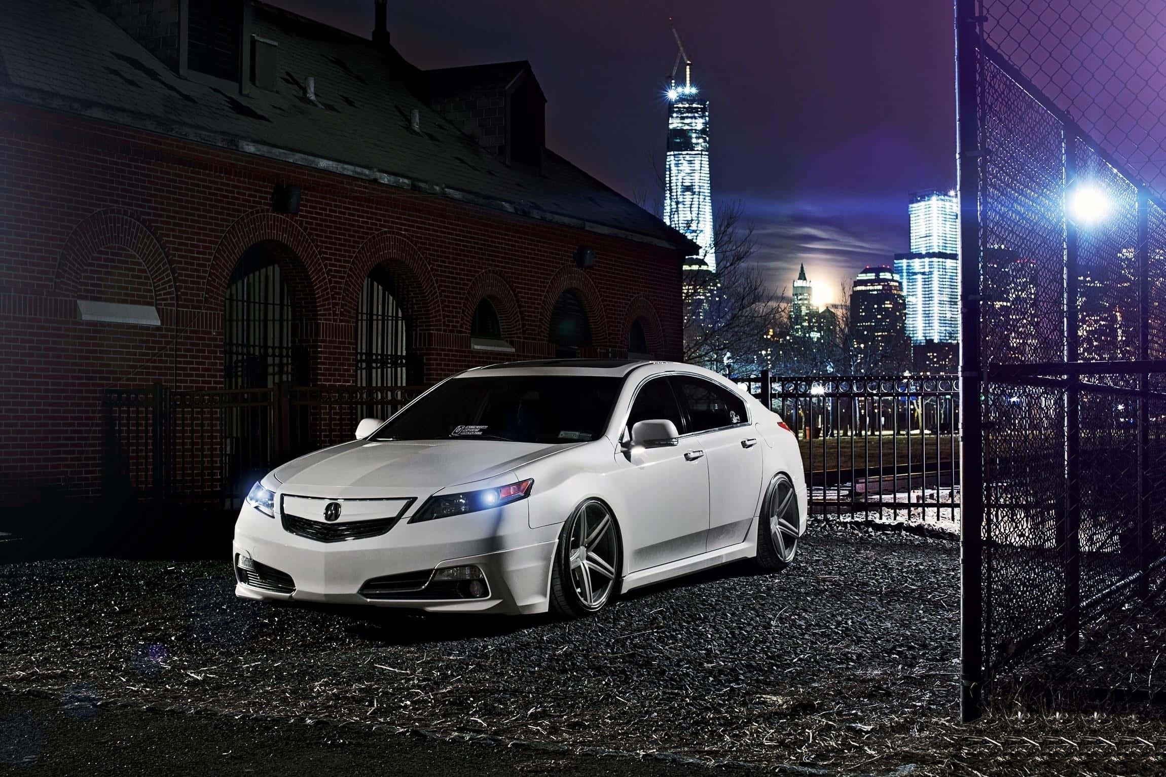 Acura Tsx 桌布 壁紙 — 一輛時尚的白色跑車在夜間停在一棟磚砌建築旁，背景是閃爍的城市天際線。