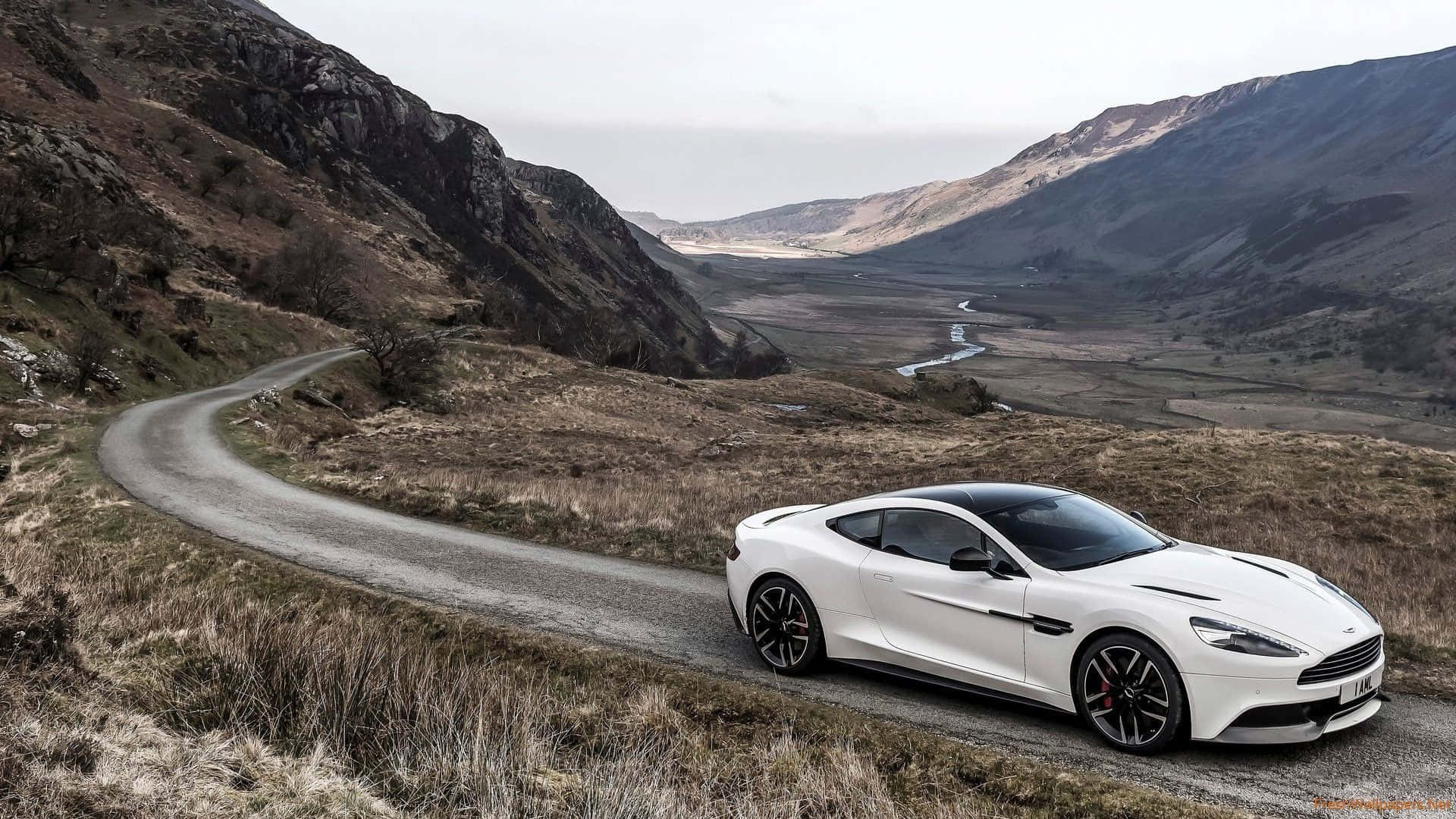 Aston Martin Vanquish 桌布 壁紙 — 一輛時尚的白色跑車在陰天下行駛在崎嶇山區蜿蜒的碎石路上。
