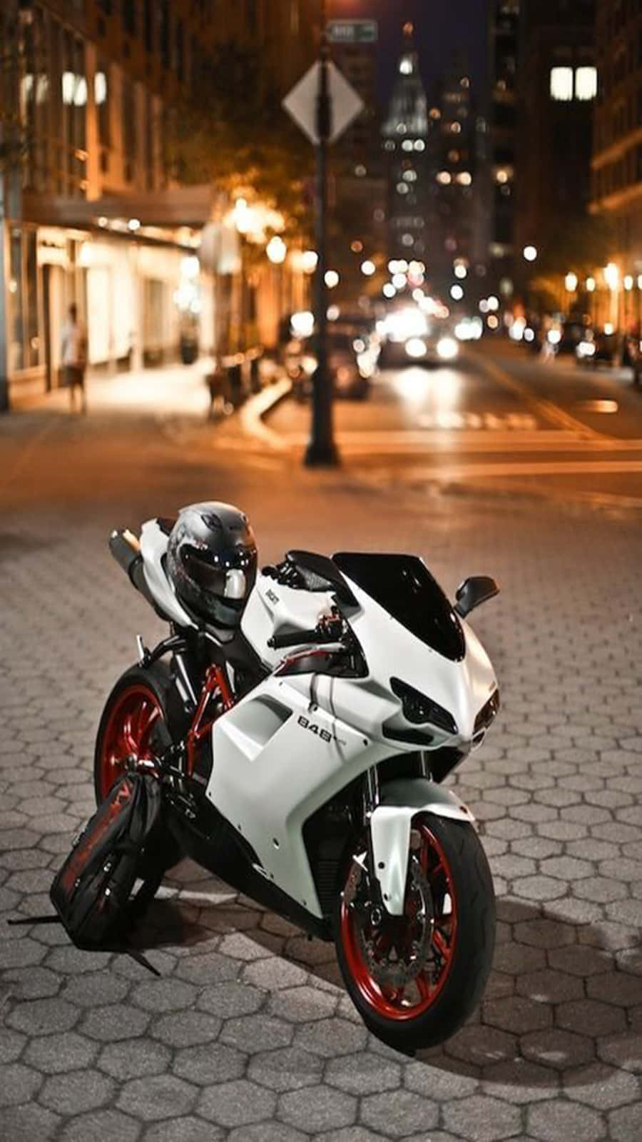  壁紙 — 一輛時尚的白色運動機車，紅色輪胎，夜晚停泊在鵝卵石街道上，城市燈光下。