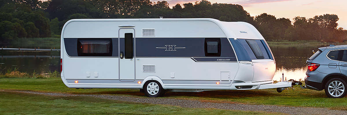 Caravan 桌布 壁紙 — 一輛時尚的白色露營車帶有深藍色點綴，停在湖邊，夕陽西下，連接著一輛銀色SUV。