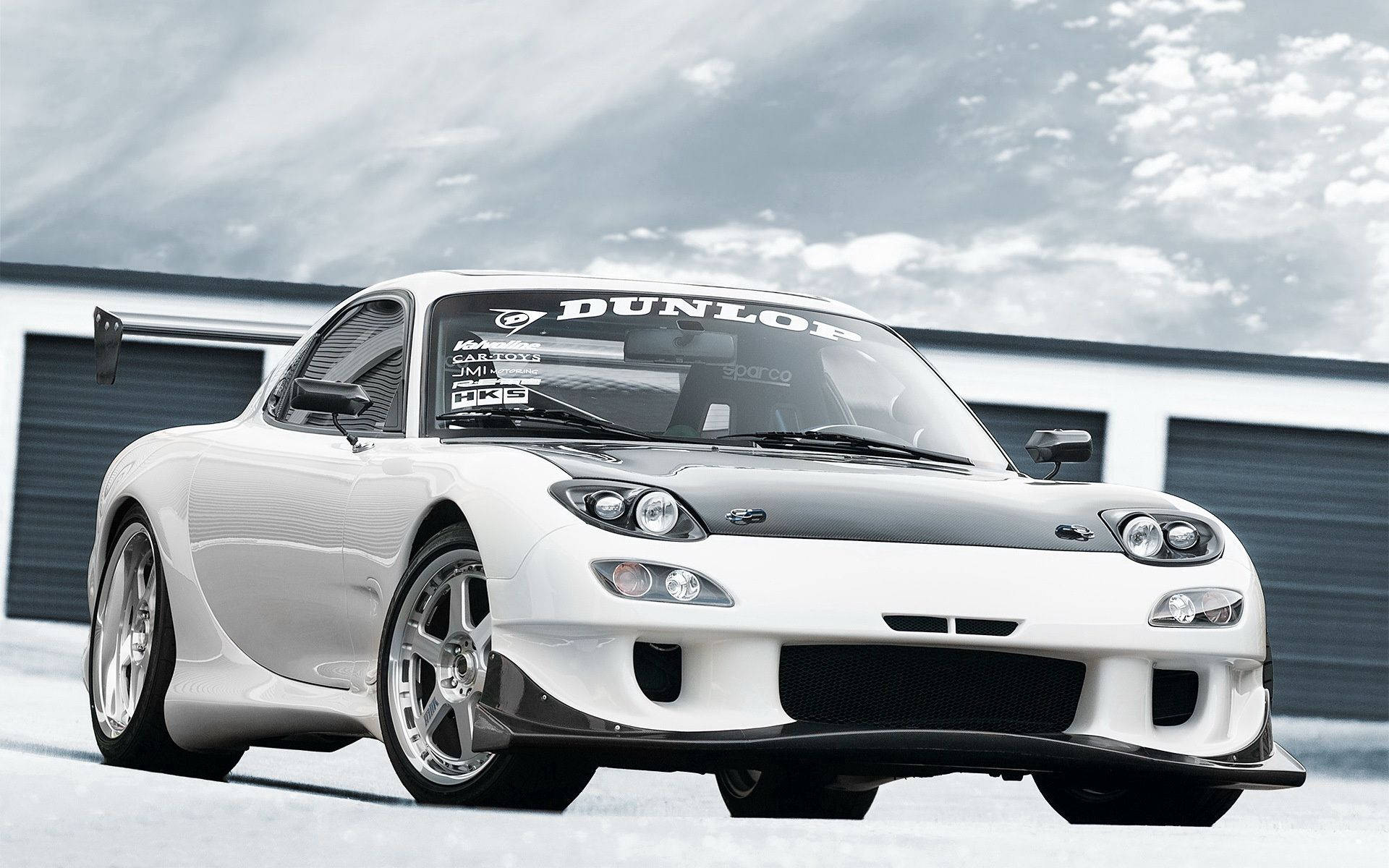 Mazda 桌布 壁紙 — 一輛時尚的白色馬自達RX-7跑車，帶有黑色飾邊和Dunlop貼紙，停放在車庫前，天空多雲。