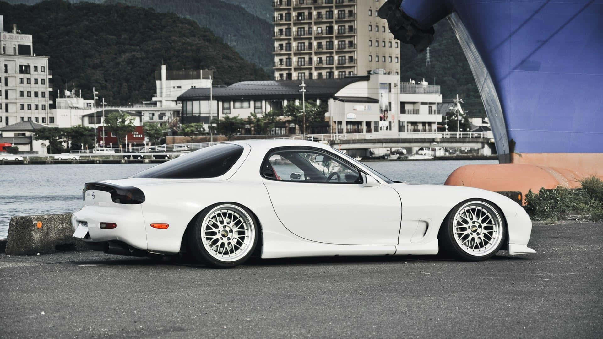 Mazda 桌布 壁紙 — 一輛時尚的白色馬自達RX-7跑車停泊在海濱，背景有城市建築物和山丘。