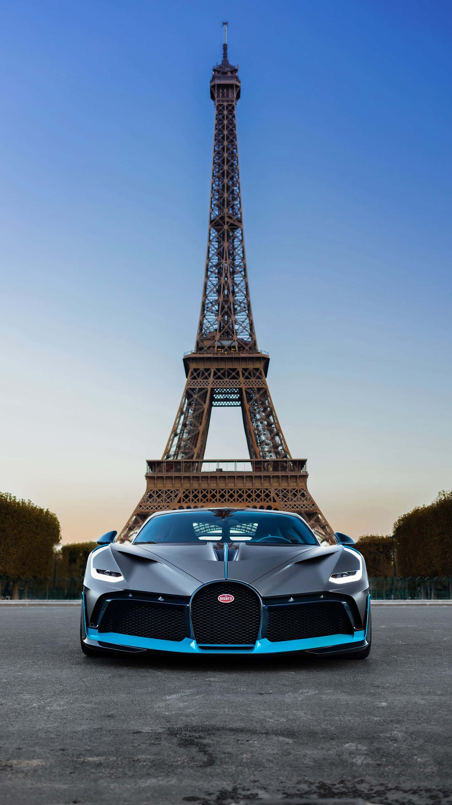 Bugatti Phone 桌布 壁紙 — 一輛時尚的磨砂灰色布加迪帶有鮮豔的藍色點綴，在黃昏時分面向著鋪砌的道路，矗立在雄偉的艾菲爾鐵塔下。
